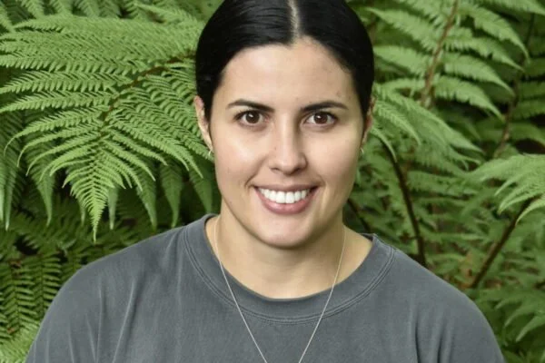 A woman with dark hair smiling, wearing a gray shirt and a necklace, standing outdoors in front of green ferns.