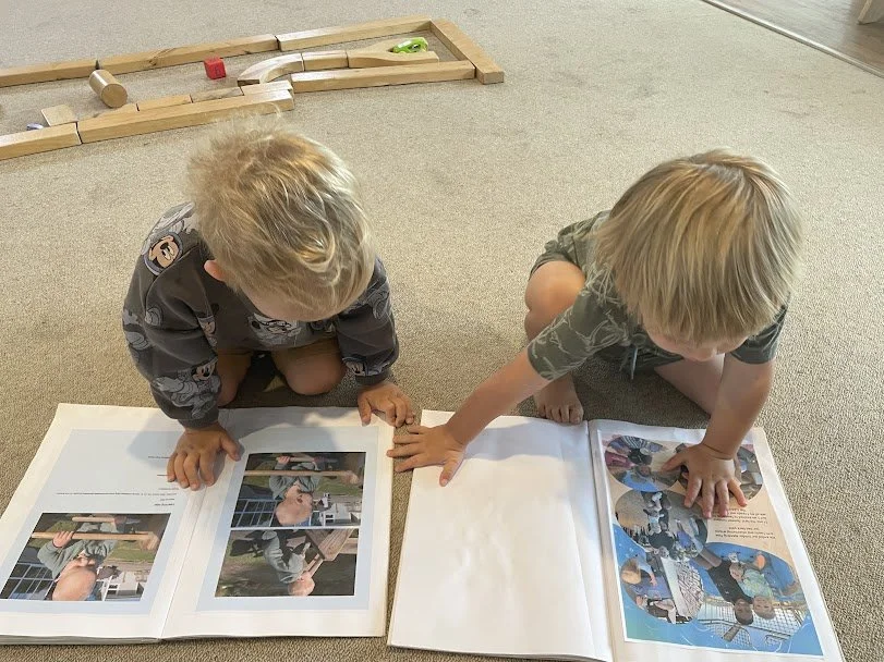 Two young children sit on a carpeted floor looking at a photo album. One child points to the pictures while the other looks on. A wooden train track with toy trains and other toys is in the background.