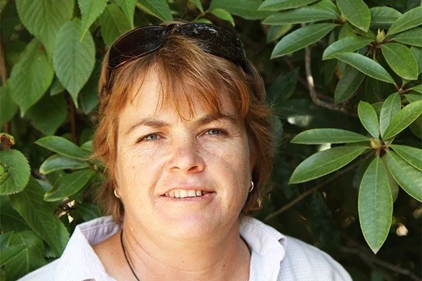 A woman with short light brown hair and sunglasses on her head, smiling, standing in front of green bushy foliage.