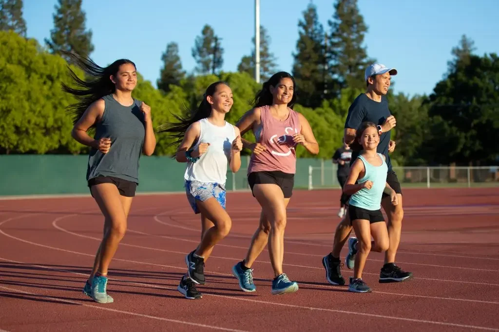 A group of five people, two women, a man, and two young girls, running on a track in an outdoor stadium with green trees in the background.