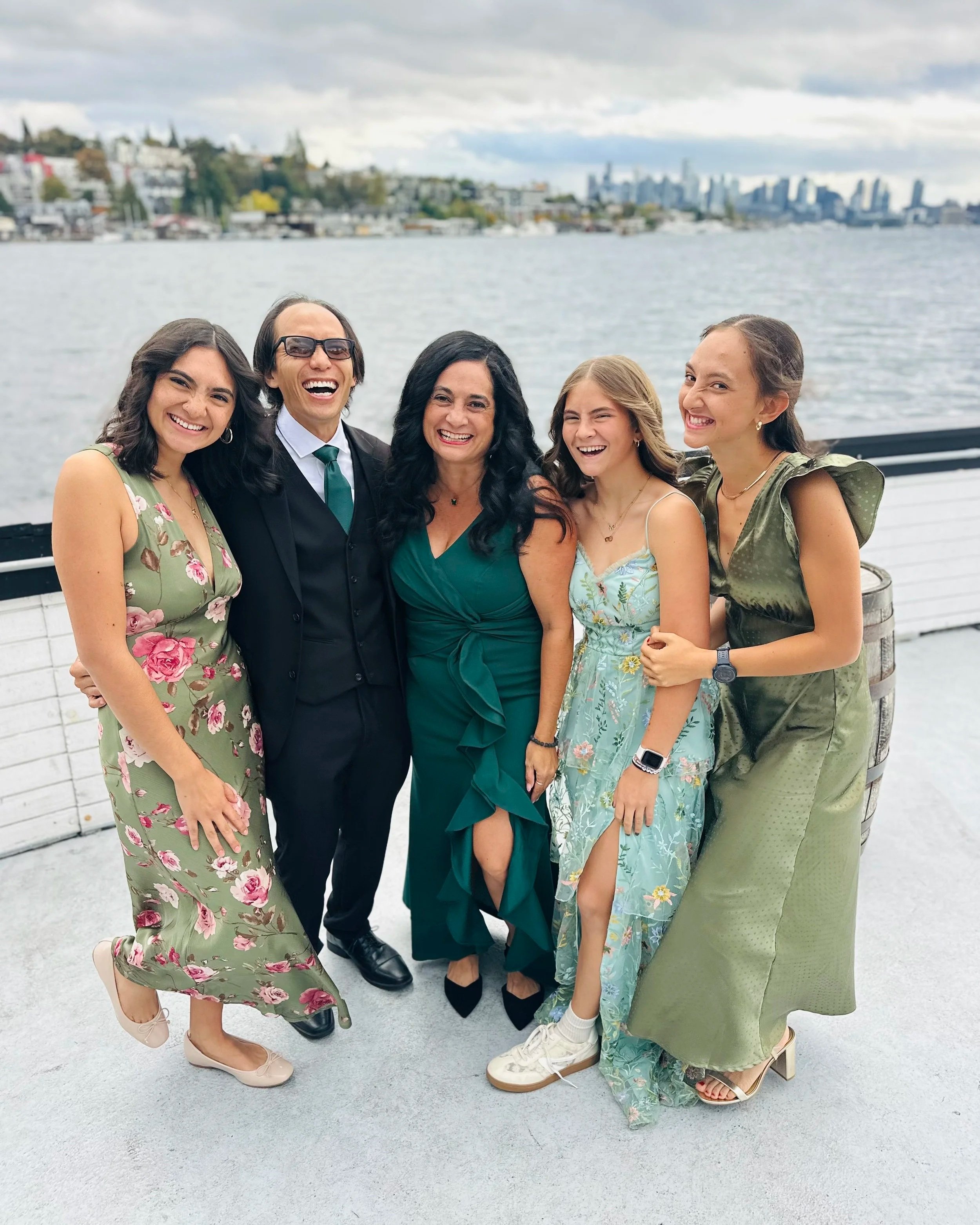 Group of four women and a man standing outdoors near water, smiling, and dressed in colorful dresses and formal wear, with a city skyline and houses in the background.