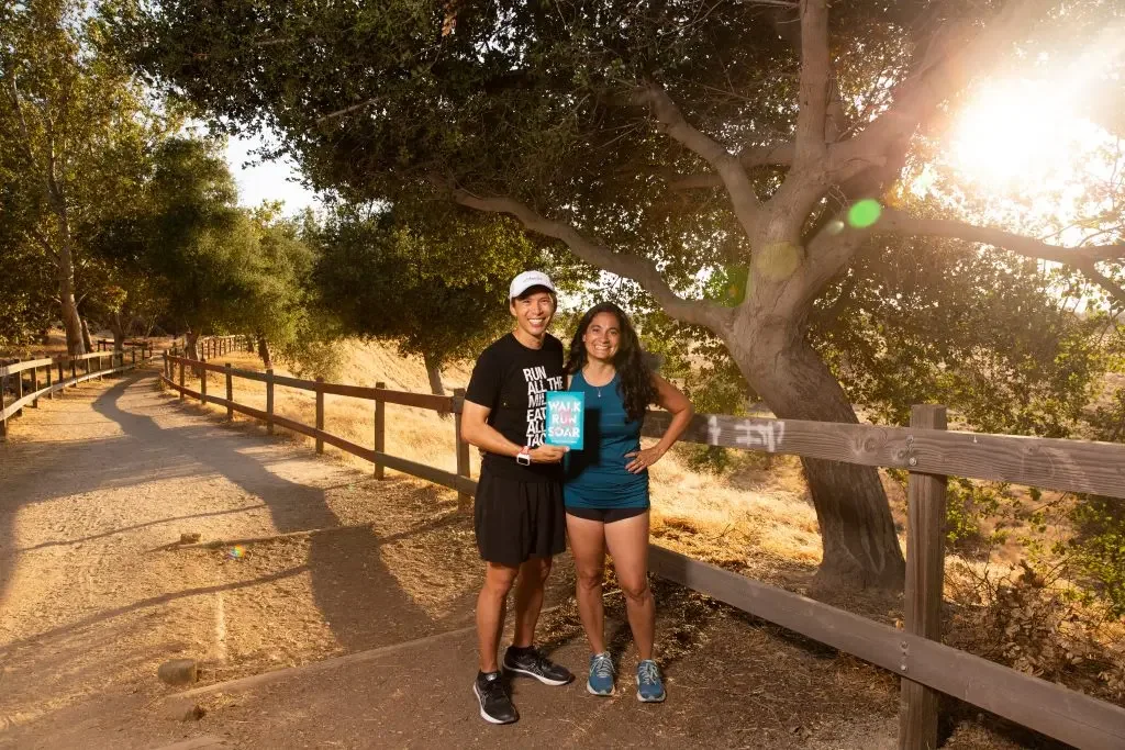 Two women standing on a sunlit hiking trail with a large tree in the background, one holding a book and both smiling.
