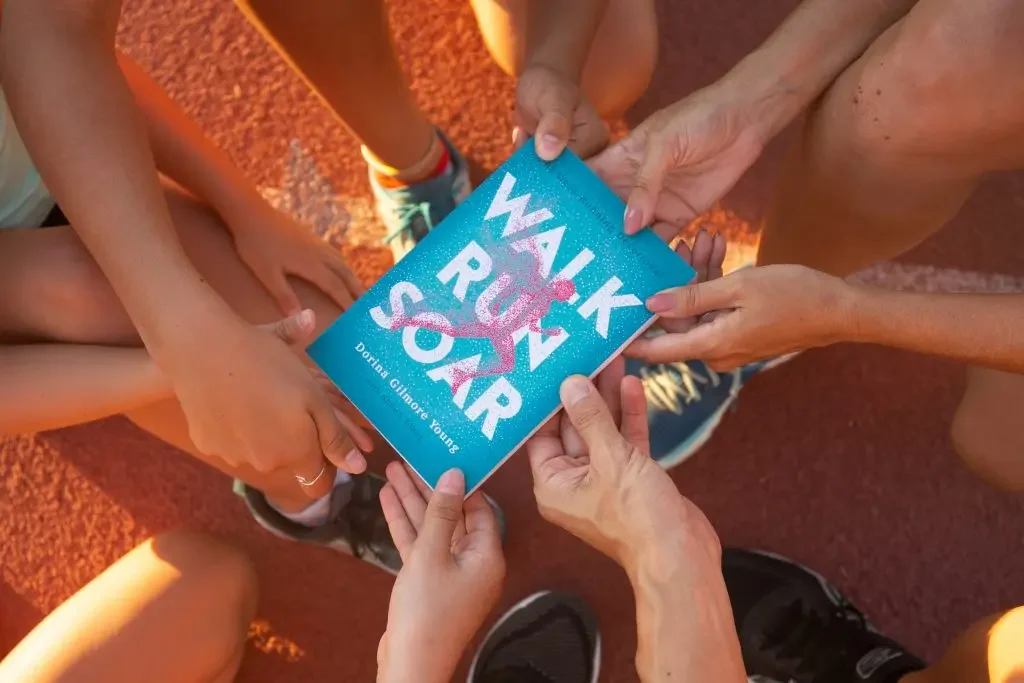 Multiple people holding a blue book titled 'WALK RUN SOAR' on a red track surface, with a young girl in running shoes in the background.
