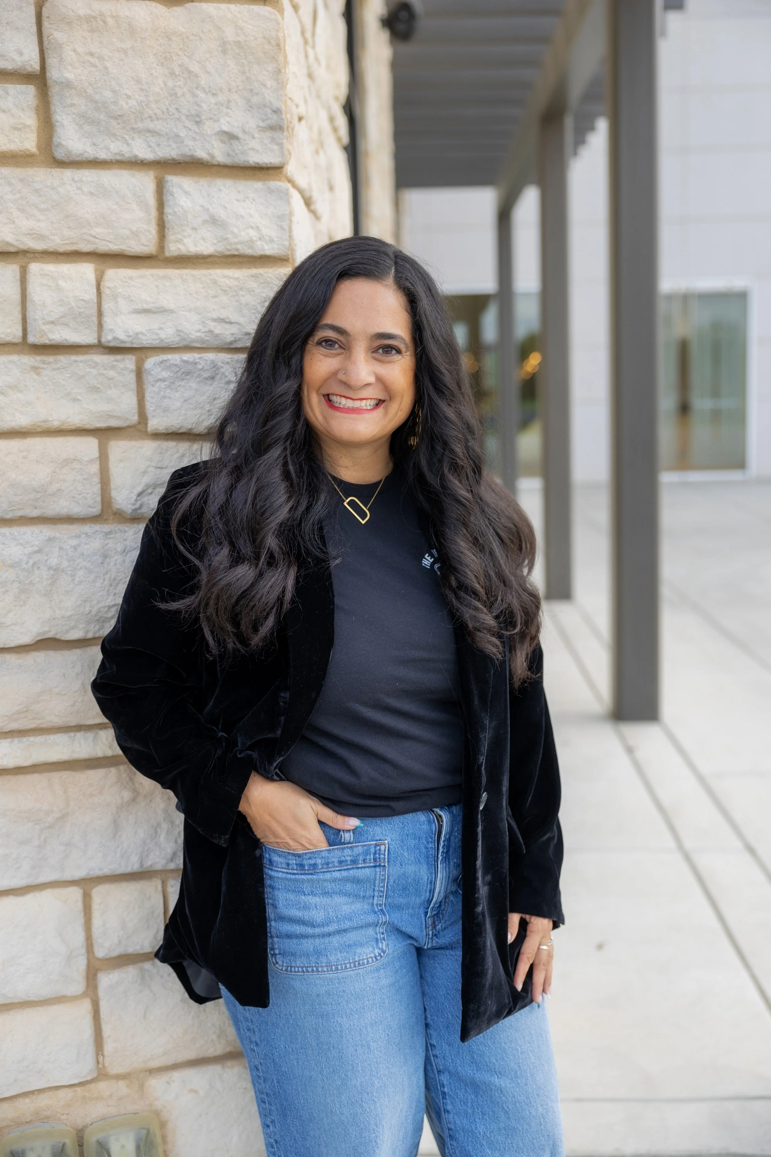 Smiling woman with long black hair, wearing a black velvet jacket, black T-shirt, and jeans, standing against a beige brick wall outside.