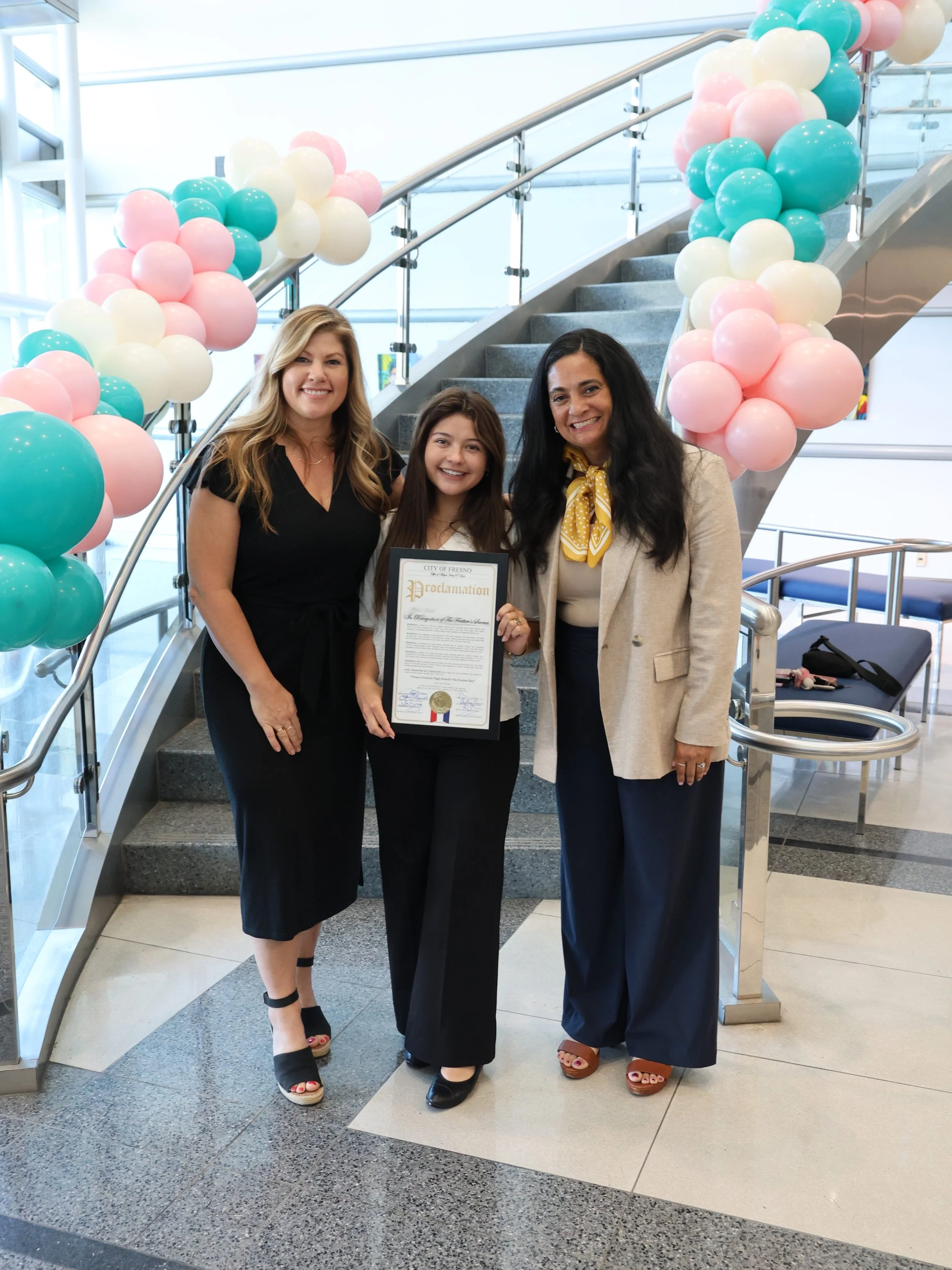 Three women standing on stairs decorated with pastel pink, white, and teal balloons, one woman holding a certificate. They are smiling, dressed in business casual attire, in an indoor setting with modern design.