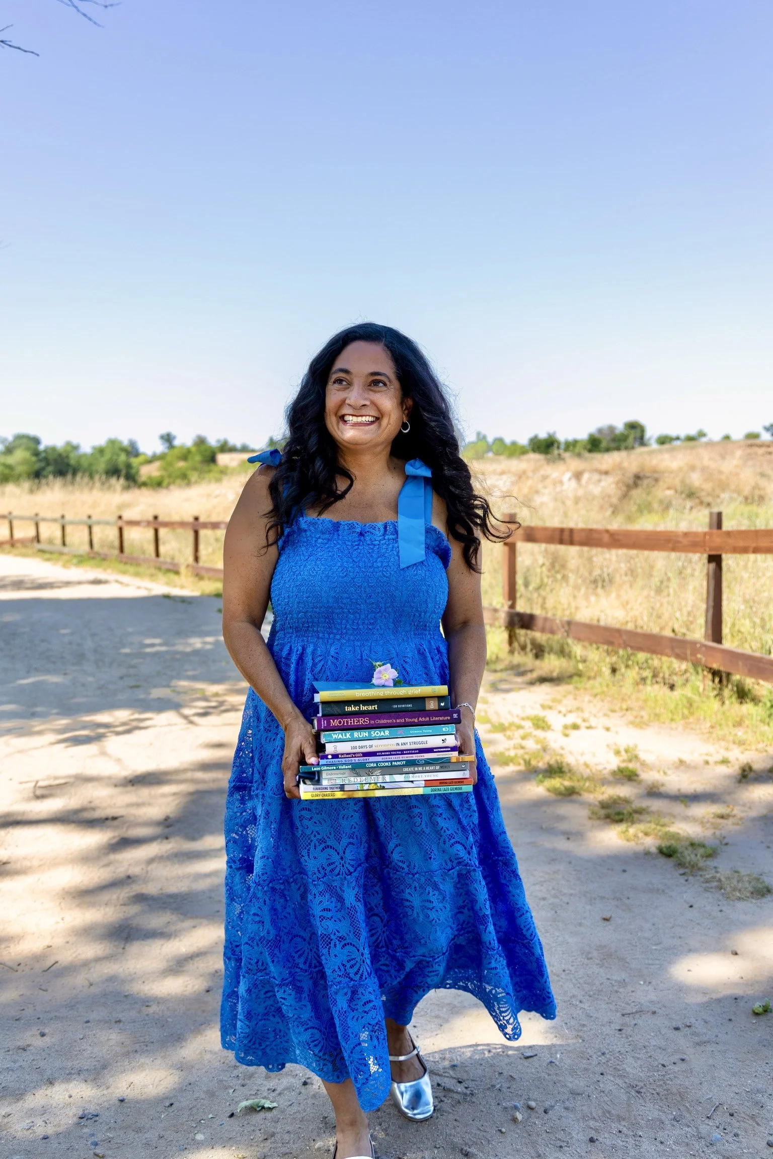 A woman in a blue lace dress holding a stack of books outdoors on a sunny day. She has long, dark, wavy hair and is smiling. The background includes a dirt path, wooden fence, dry grass, shrubs, and a clear blue sky.