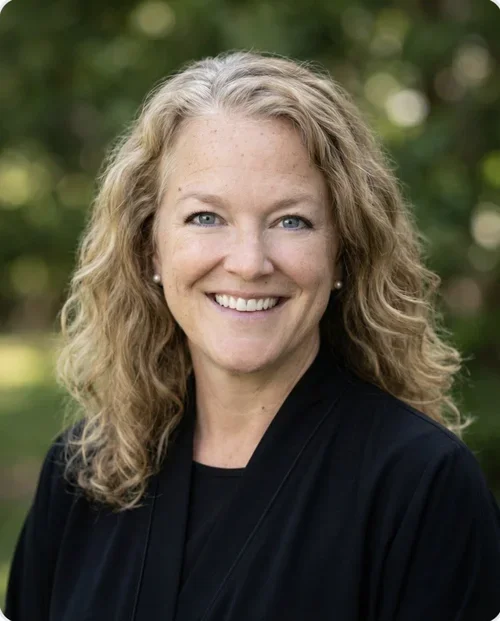 A woman with curly blonde hair, wearing a black top and pearl earrings, smiling outdoors with greenery in the background.