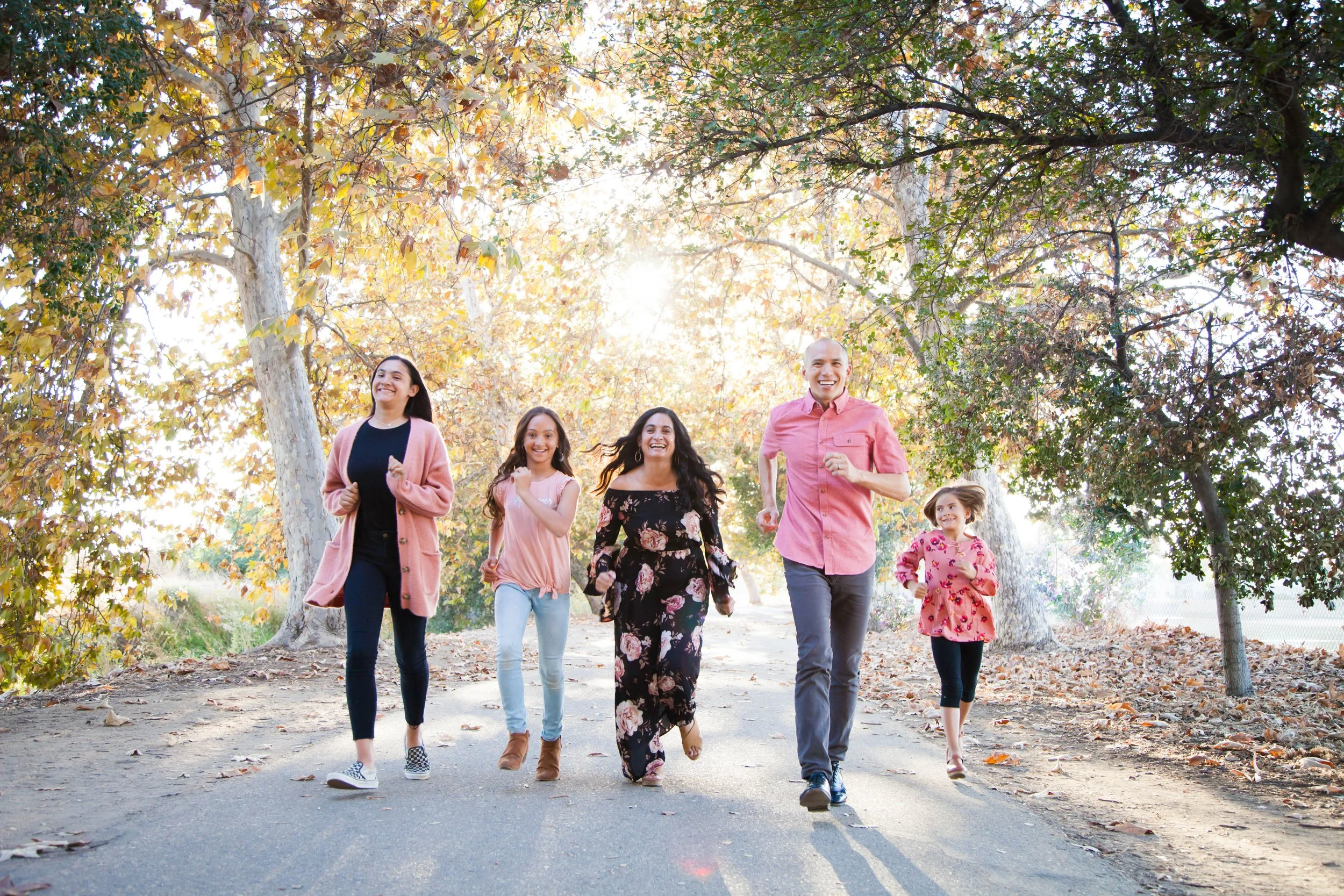 Group of five people, two adult women, one adult man, and two young girls, walking and smiling outdoors under trees with autumn leaves.