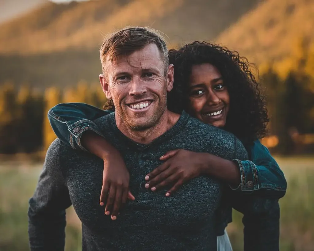 A smiling man giving a piggyback ride to a girl outdoors during sunset, with a blurred nature background.