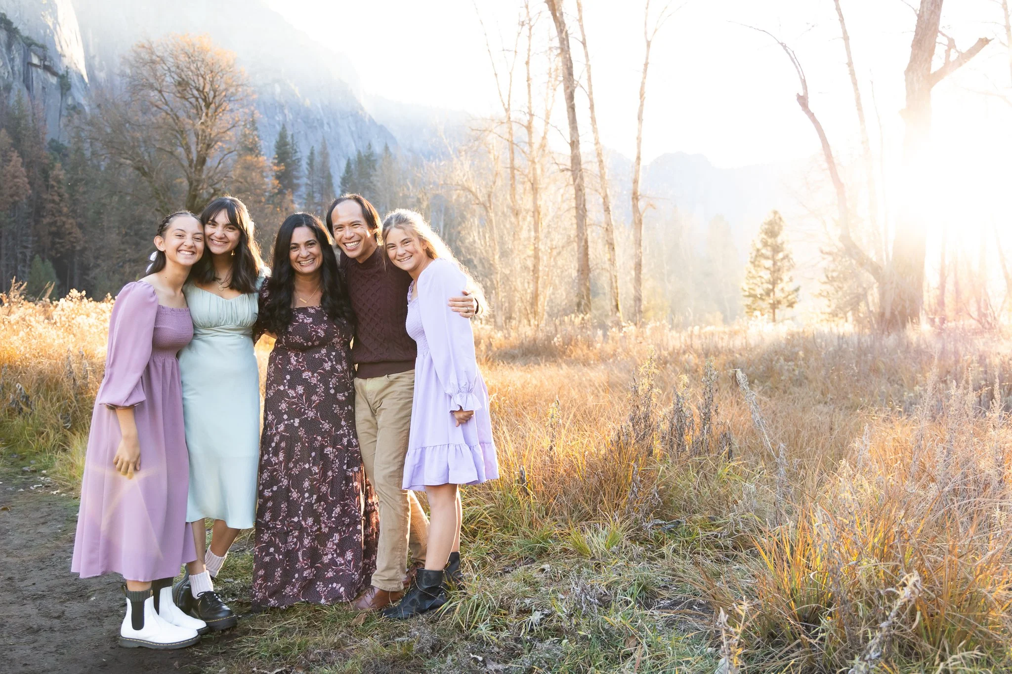A group of five smiling people, including four women and a man, standing outdoors in a natural setting with trees and mountains, bathed in warm, golden sunlight.