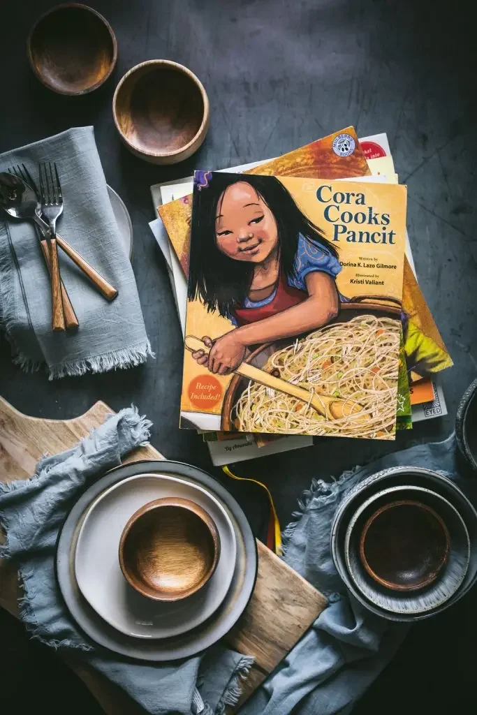 A table setting with plates, bowls, a napkin with utensils, and a cookbook titled 'Cora Cooks Pancit' featuring an illustration of a girl cooking with noodles.