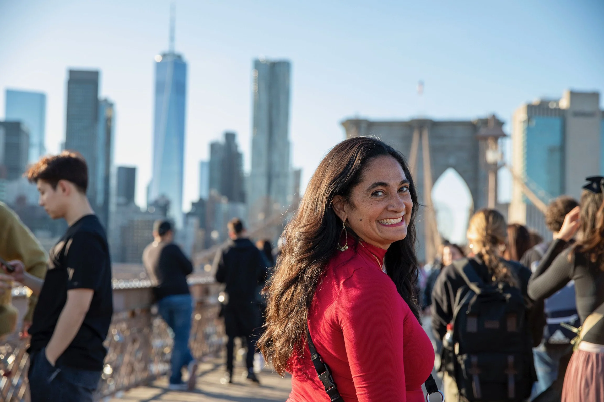 A woman in a red top smiling at the camera, with the New York City skyline and the Brooklyn Bridge in the background during the day.