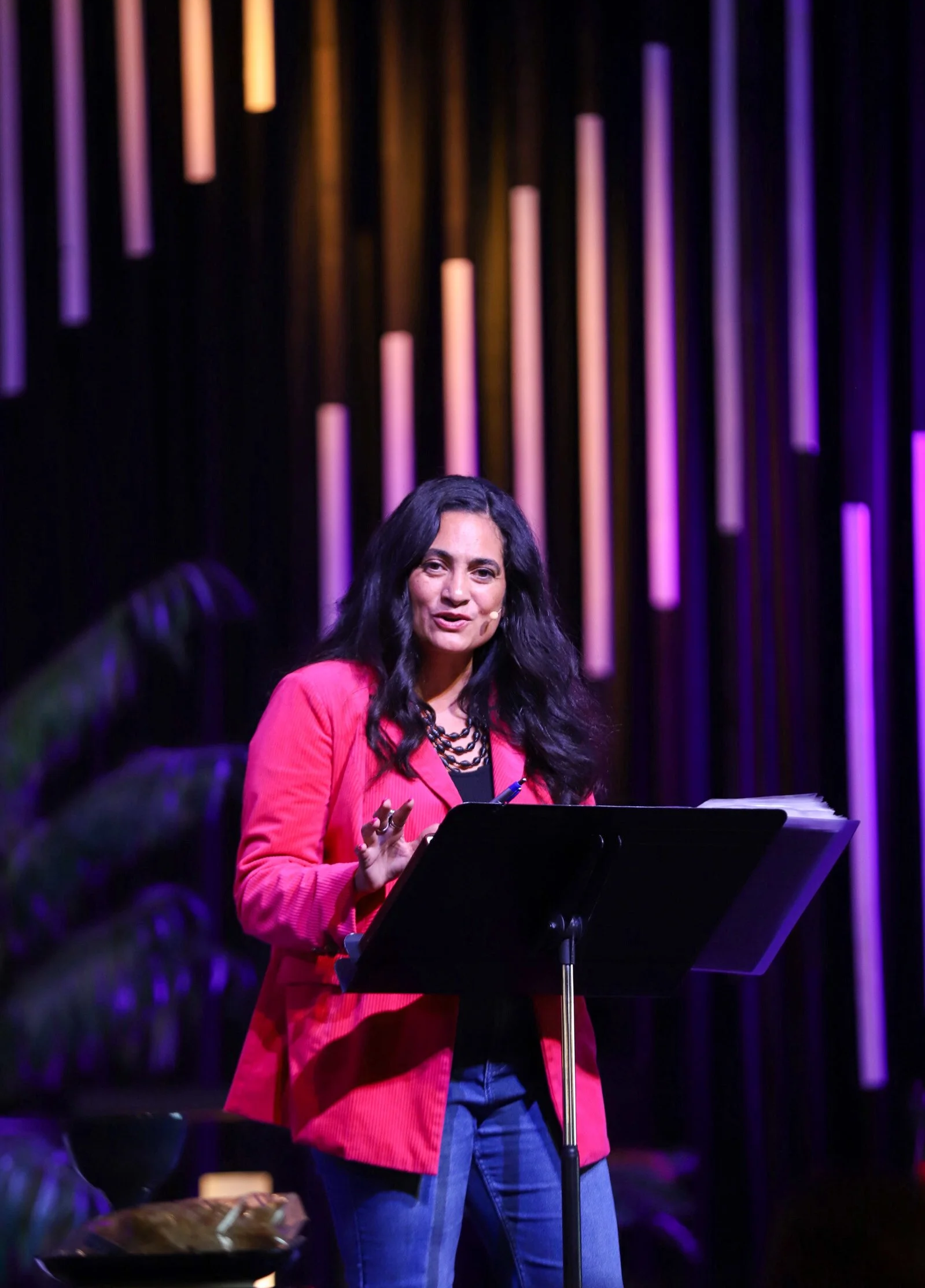 A woman with dark hair, wearing a pink blazer and blue jeans, speaking at a lectern on a stage with colorful vertical light strips in the background.