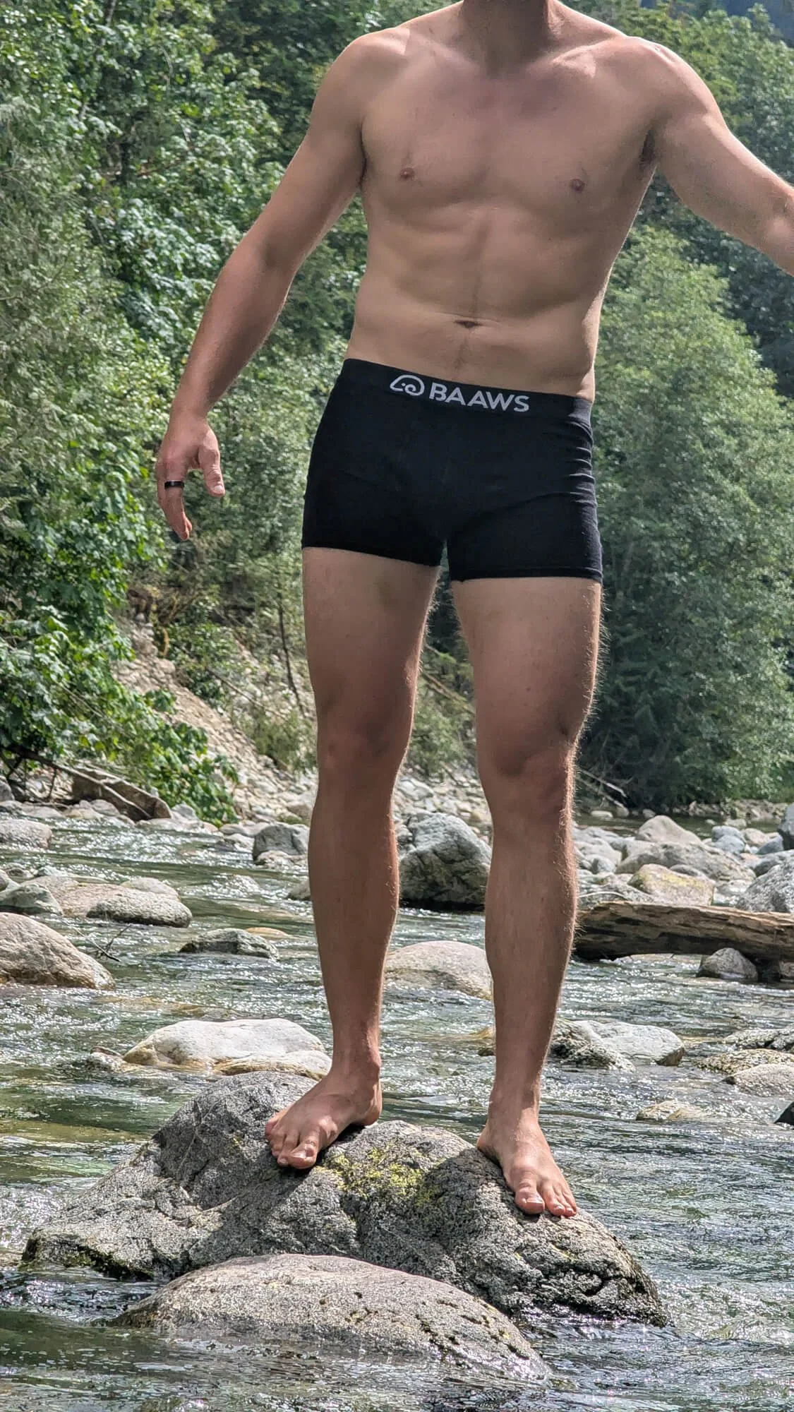 A shirtless man standing on rocks in a shallow river surrounded by green trees, wearing black shorts with the brand name "BAWS" on the waistband.