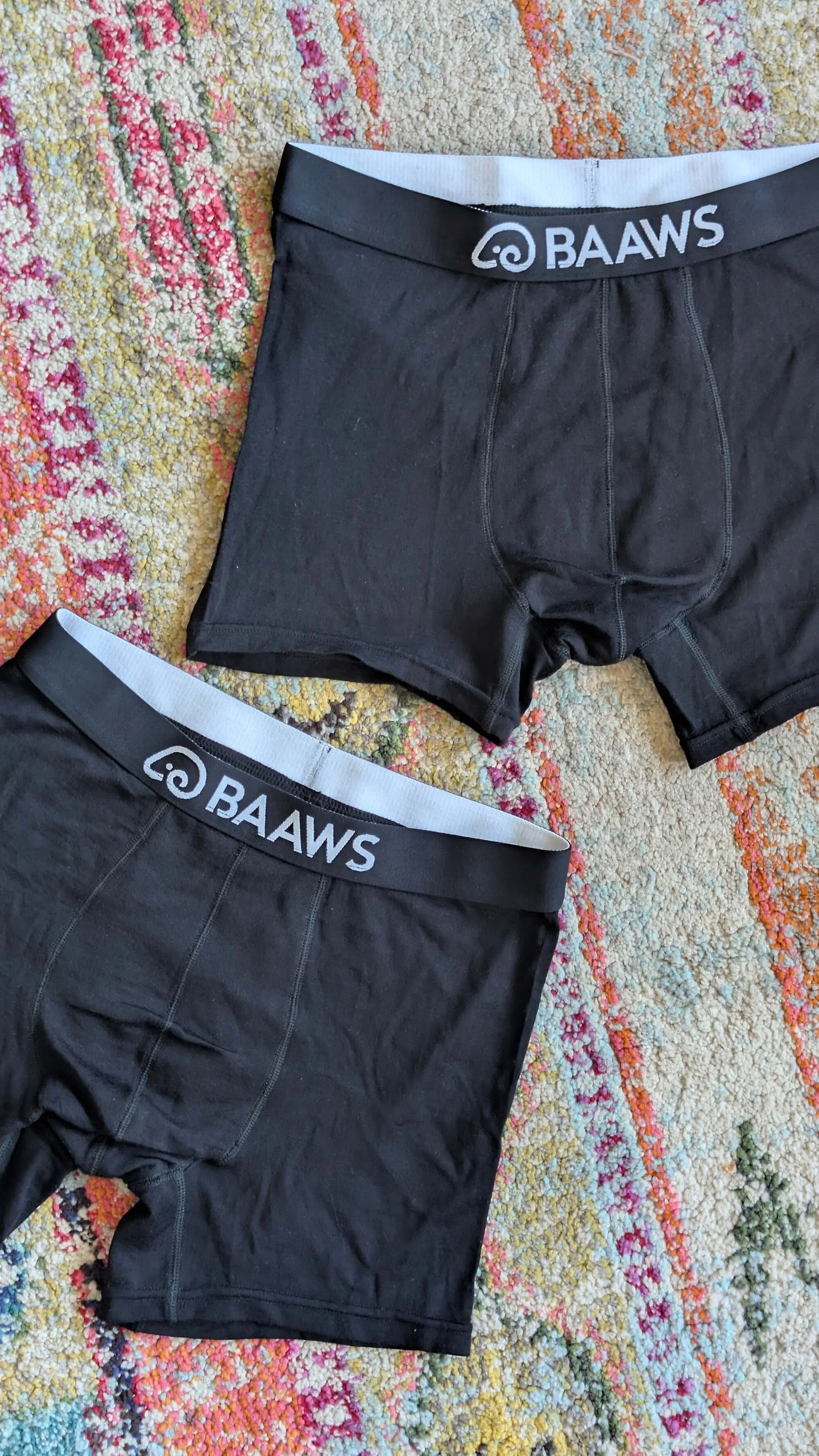 Two pairs of black boxer briefs with white waistband featuring the logo and text 'BAAWS' are lying on a colorful textured rug.