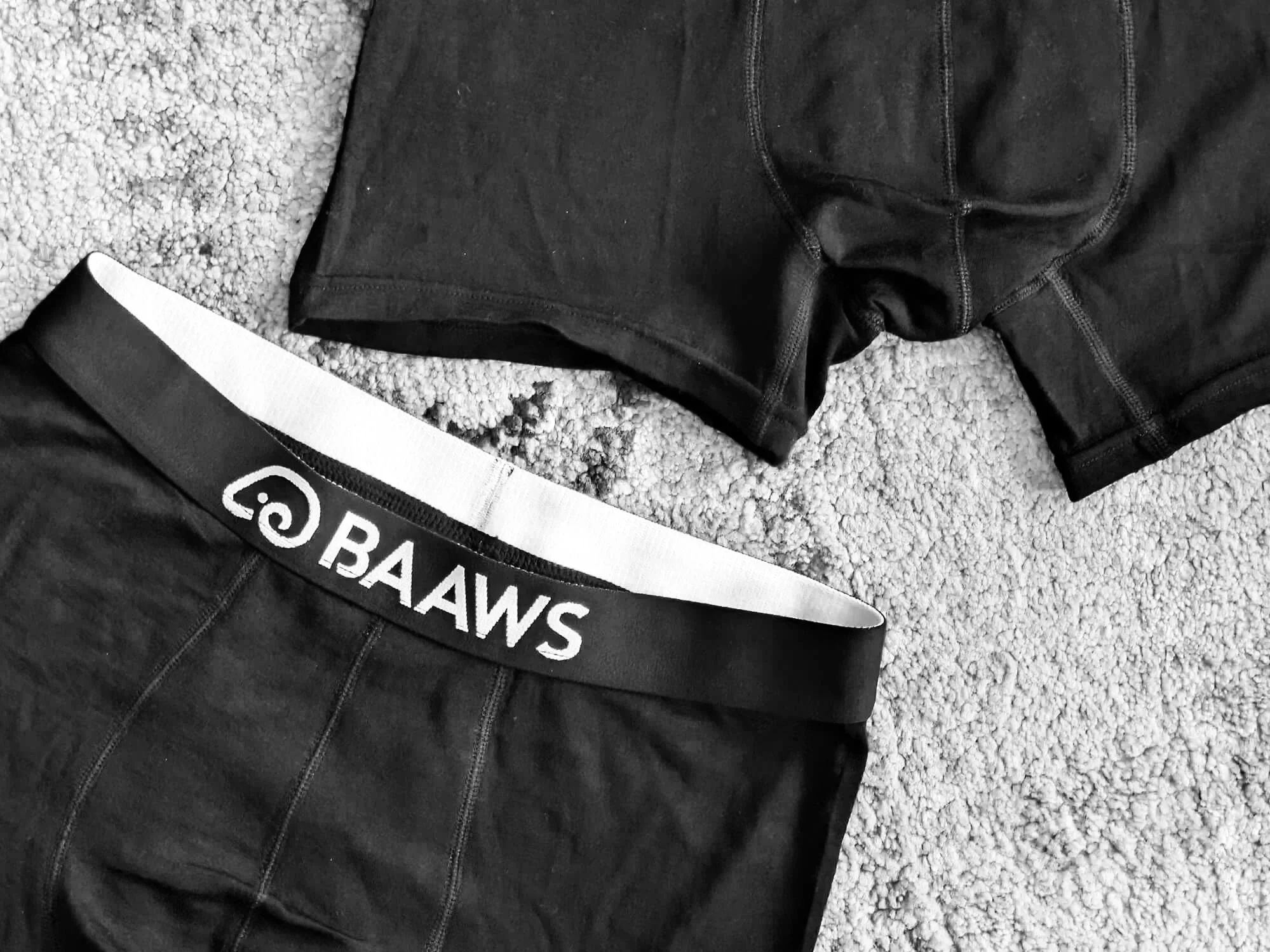 Black athletic briefs with a white waistband and black underwear with white lettering lying on a textured carpeted floor.