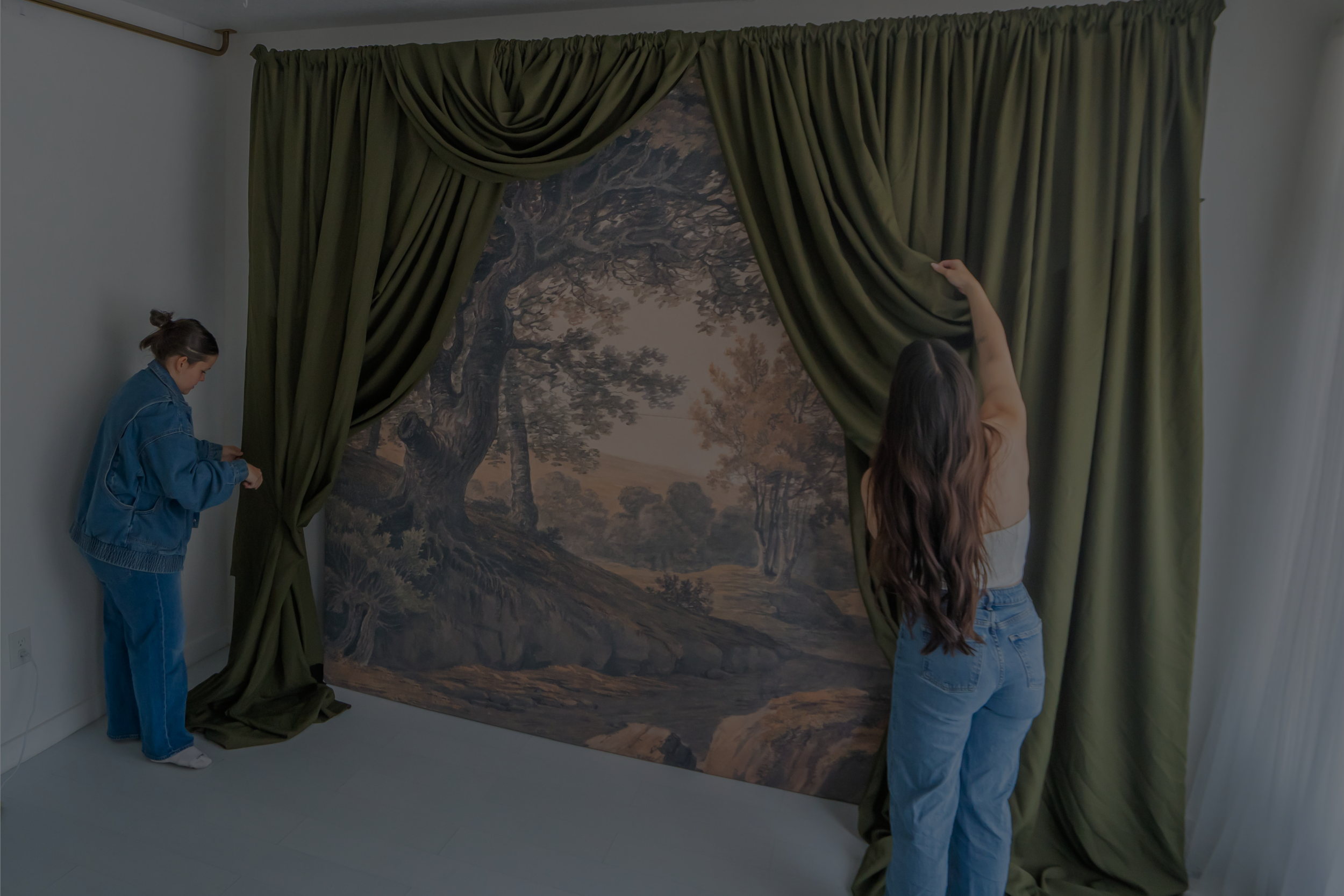 Two women are hanging a large landscape painting with trees and a cloudy sky on a blank wall. One woman is on the left side wearing a blue jacket and blue jeans, and the other woman is on the right side wearing a white top and blue jeans.