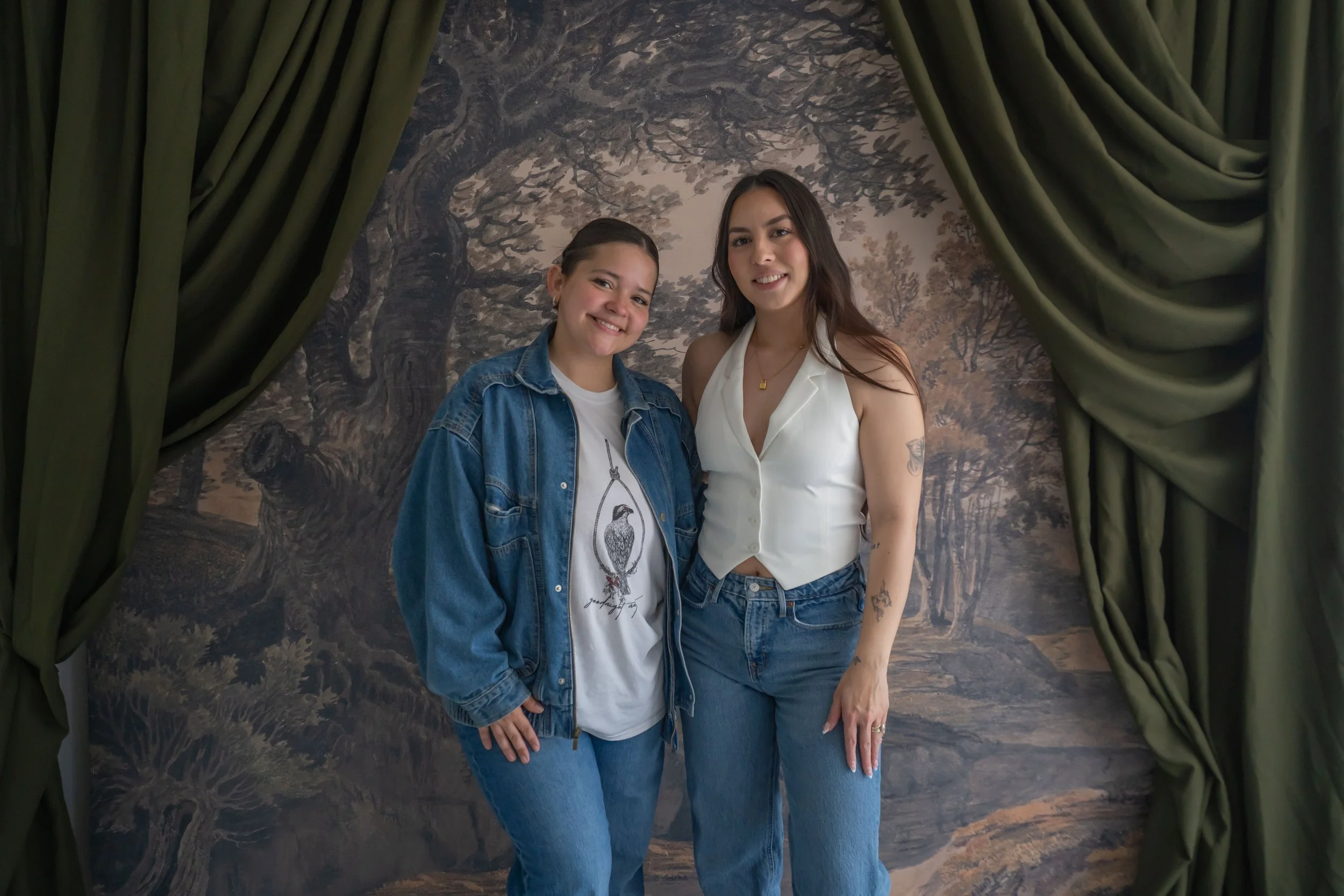 Two women standing together inside a decorative photo setup with a painted tree background, green curtains framing the scene. One woman is wearing a denim jacket and t-shirt, the other is in a white sleeveless top and jeans.
