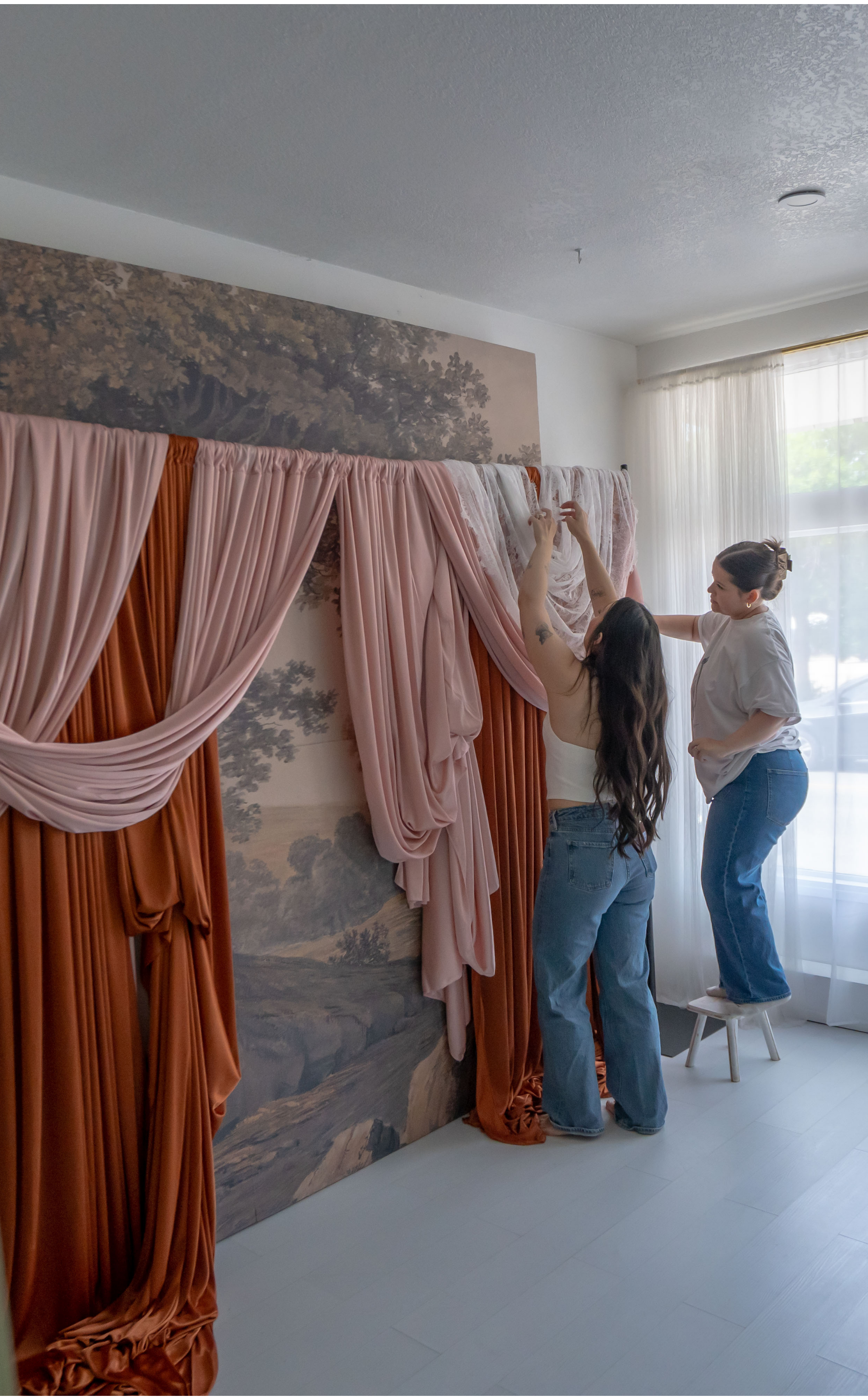 Two women are hanging pink and white drapes on a large art piece in a bright room with white walls and a window with sheer curtains.