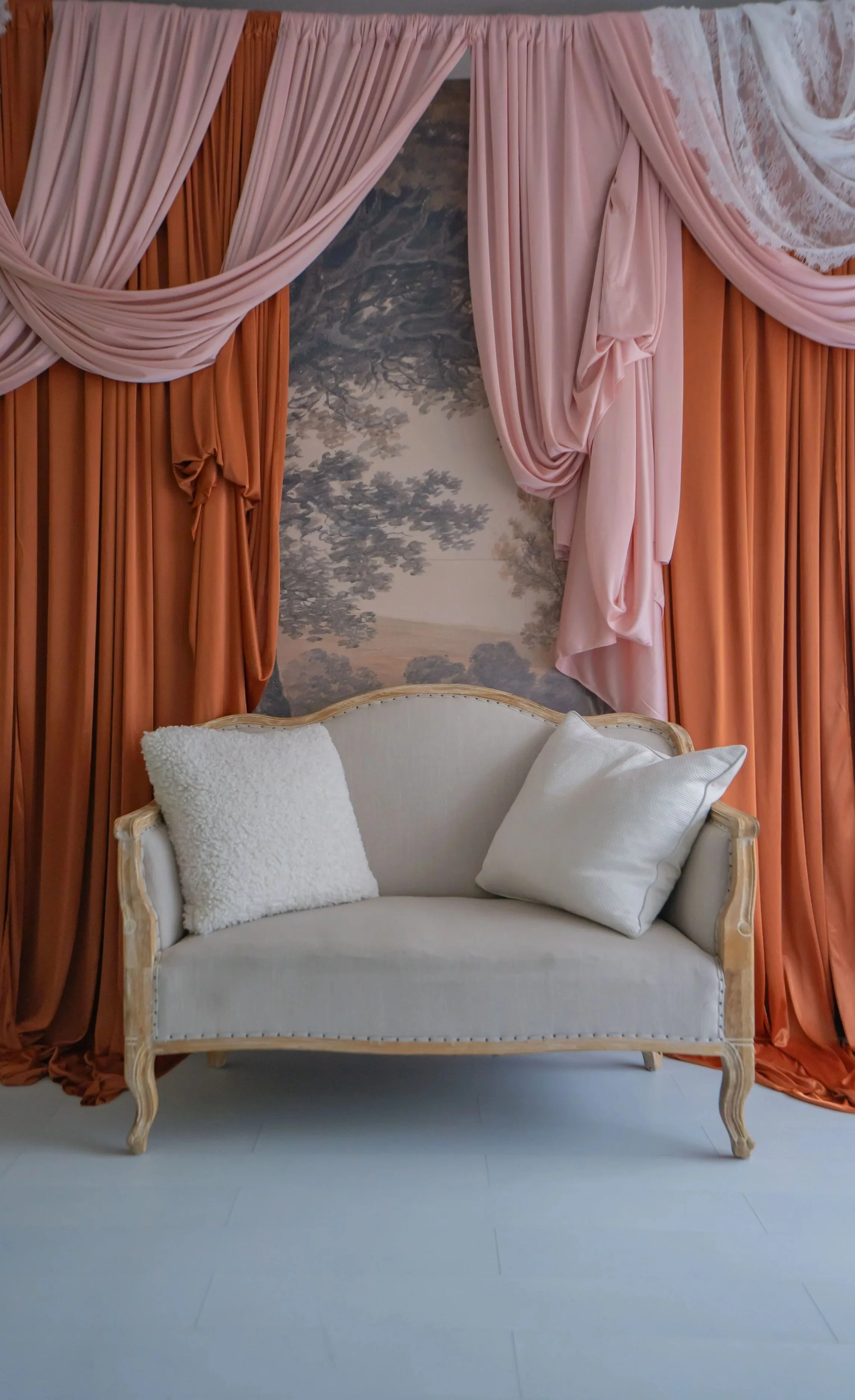 A vintage style cream-colored loveseat with ornate wooden frame and white cushions, set in front of peach and pink draped curtains and a painted scenic backdrop of trees.