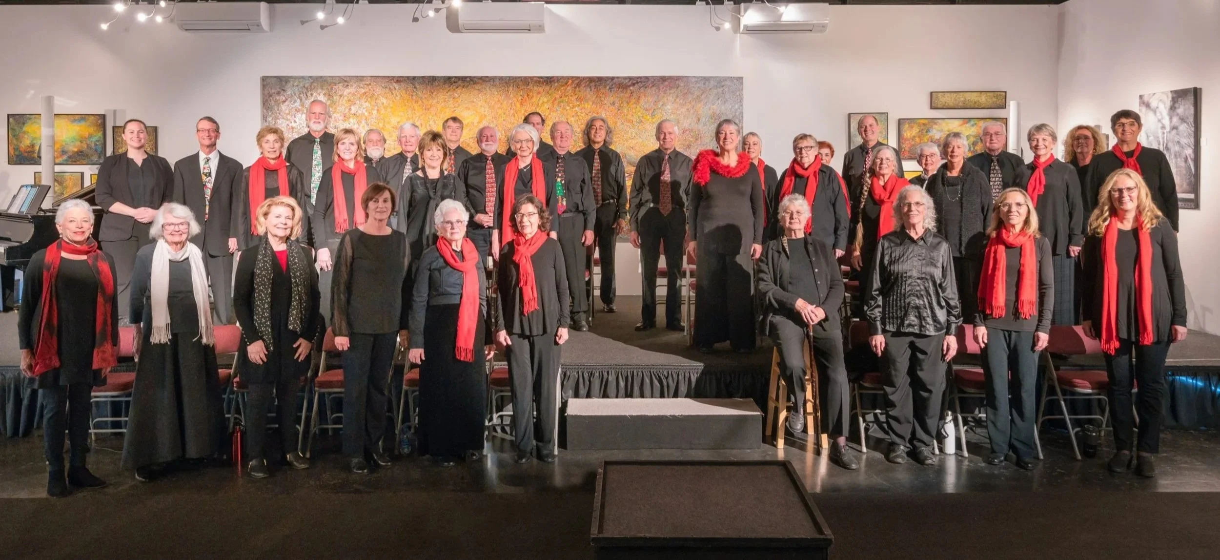 Group photo of choir members, mostly women, standing on a stage in an art gallery, many wearing black with red scarves, in front of colorful abstract paintings.