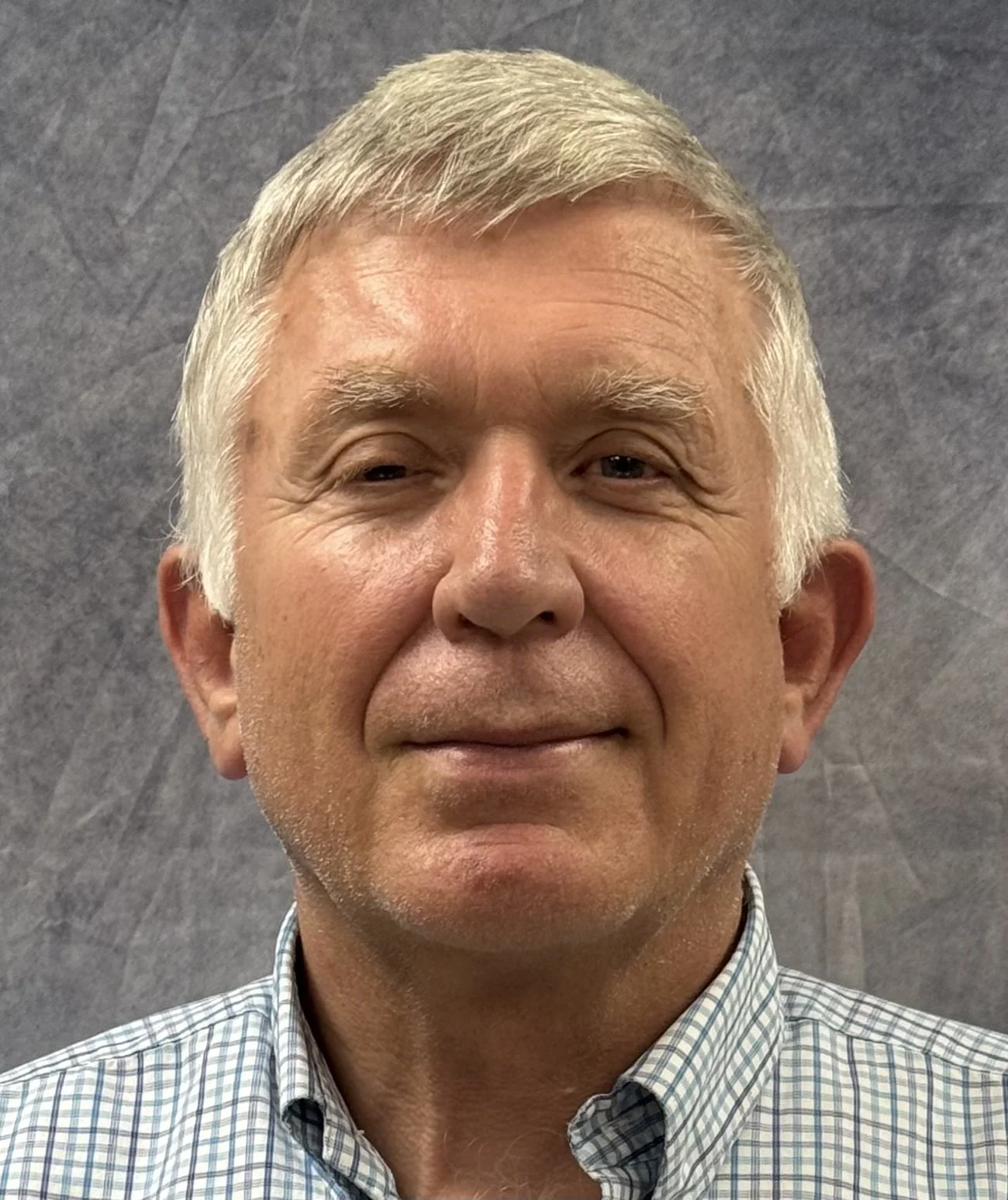 Close-up of a smiling middle-aged man with gray hair and light skin, wearing a checkered shirt, standing against a gray background.