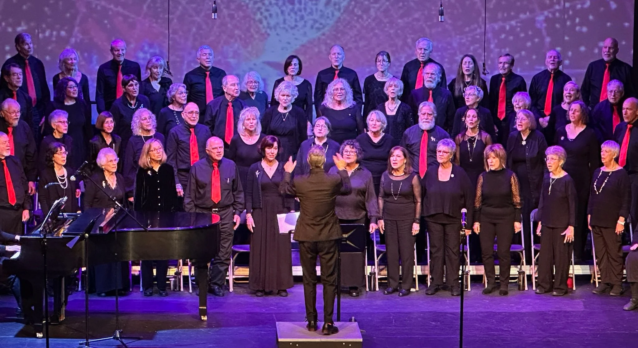 A choir performing on stage with the conductor leading them. The choir members are dressed in black, with many wearing red ties or accessories. The stage has a piano and microphones, with a colorful, abstract backdrop projected behind them.