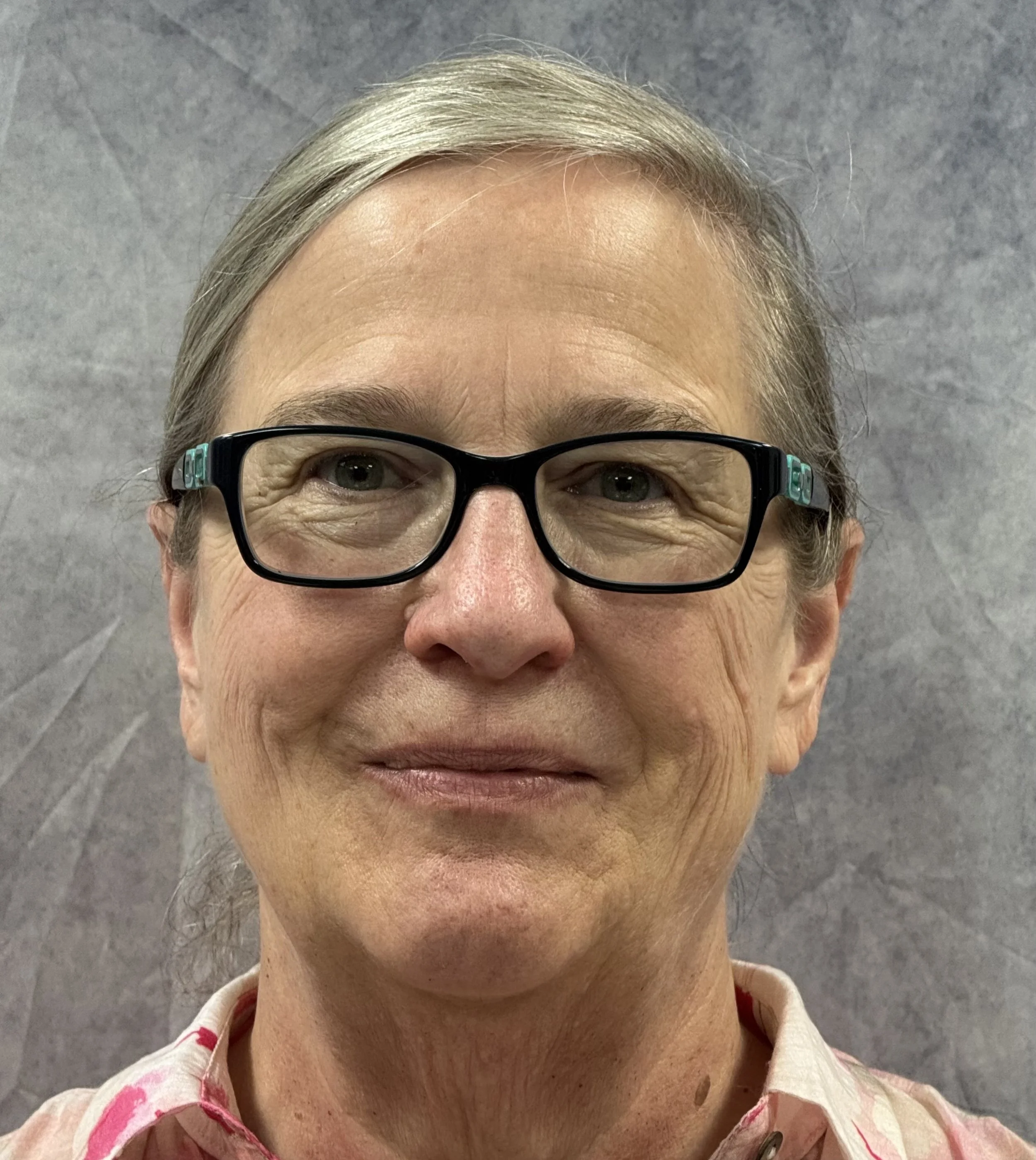 A close-up photo of a woman with short gray hair, wearing black glasses with teal accents, and a floral-patterned shirt. She is smiling slightly, with a neutral gray background.