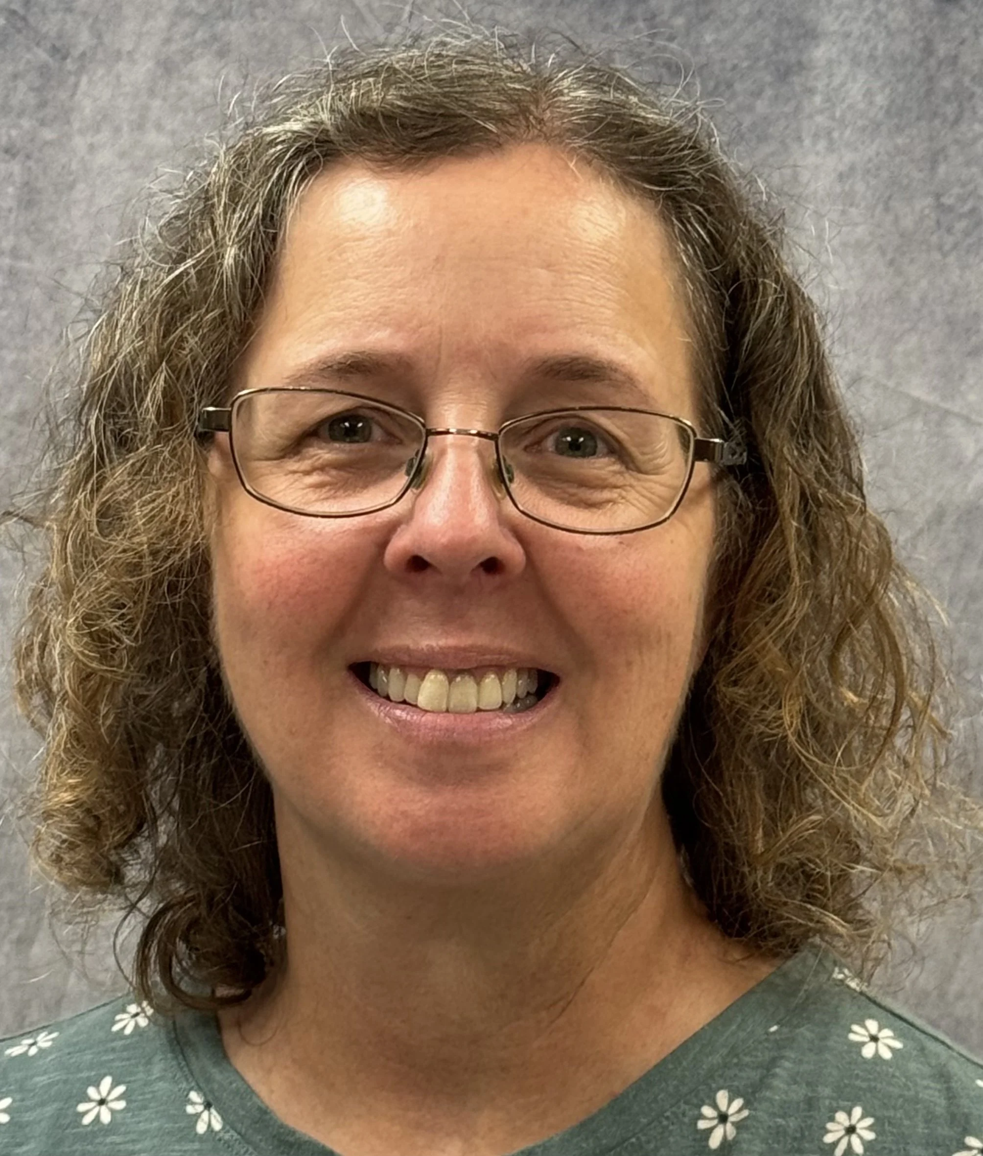 Close-up of a middle-aged woman with curly brown hair, glasses, and a wide smile, wearing a green top with white floral pattern, standing against a gray background.