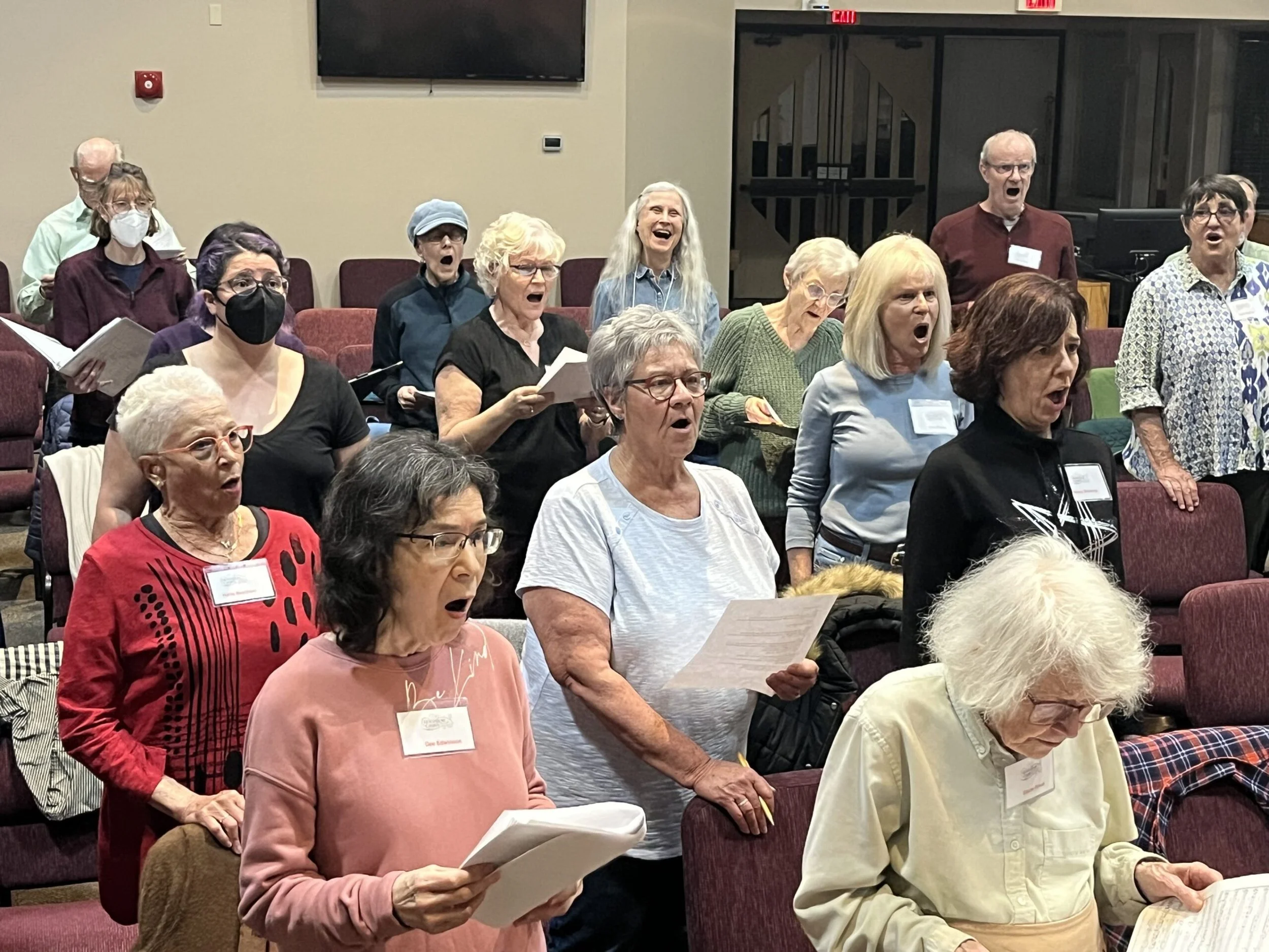 A group of mostly elderly people singing or reading from papers while standing in a conference room, some wearing glasses, masks, and name badges.