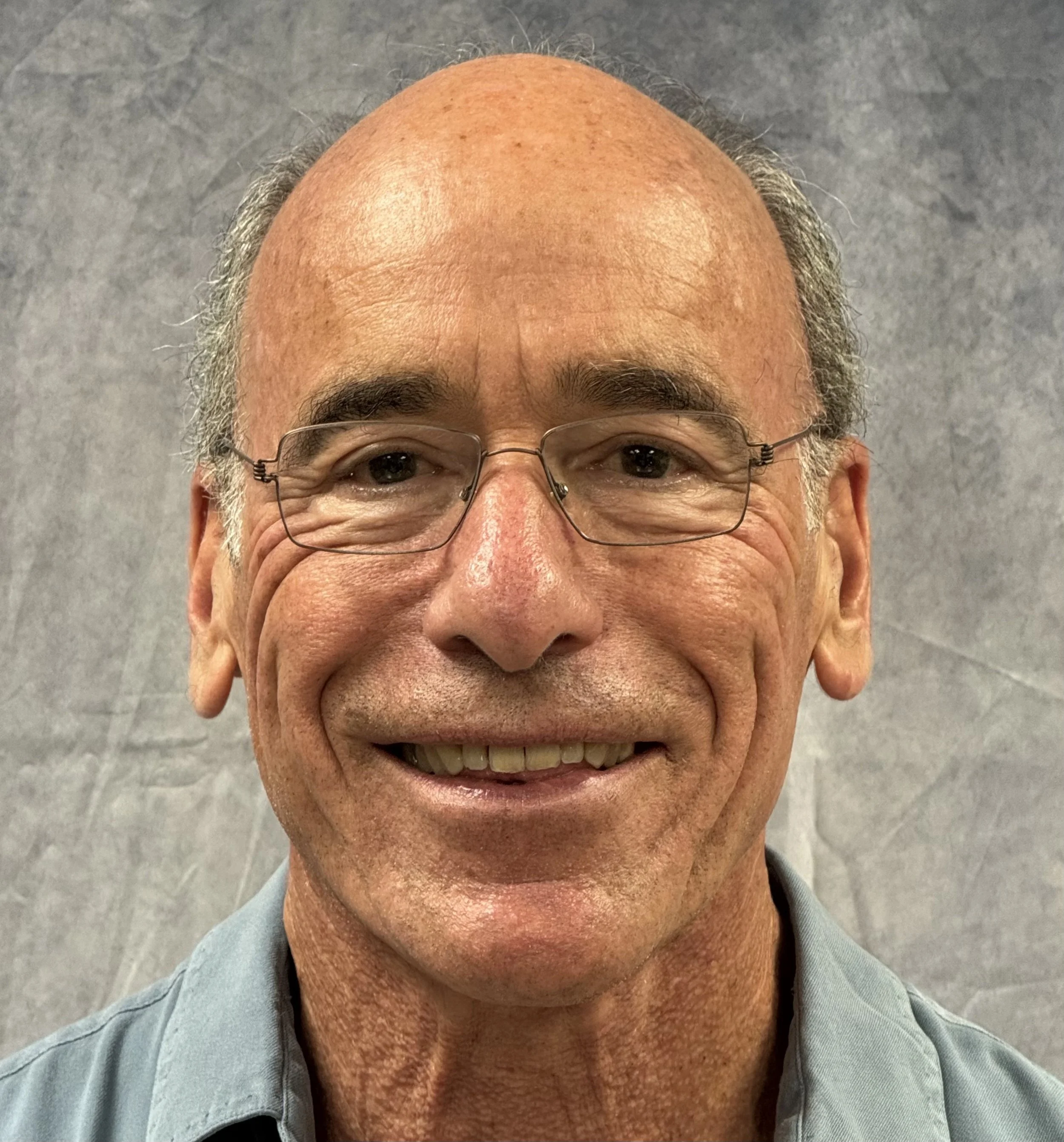 A close-up of an elderly man's face, wearing glasses and smiling, with a gray background.