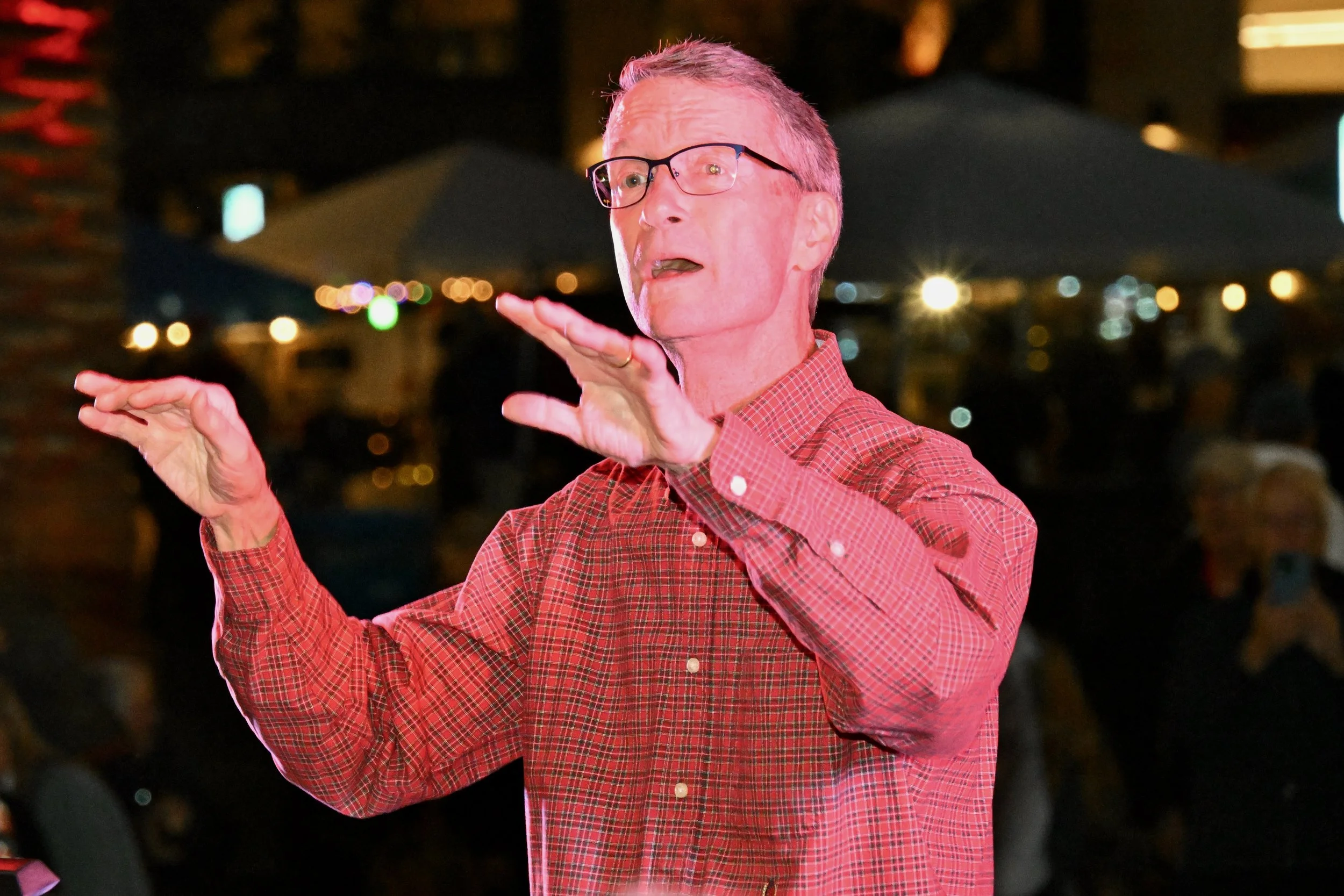 A man wearing glasses and a red checkered shirt gesturing with his hands during a night event with blurred city lights in the background.