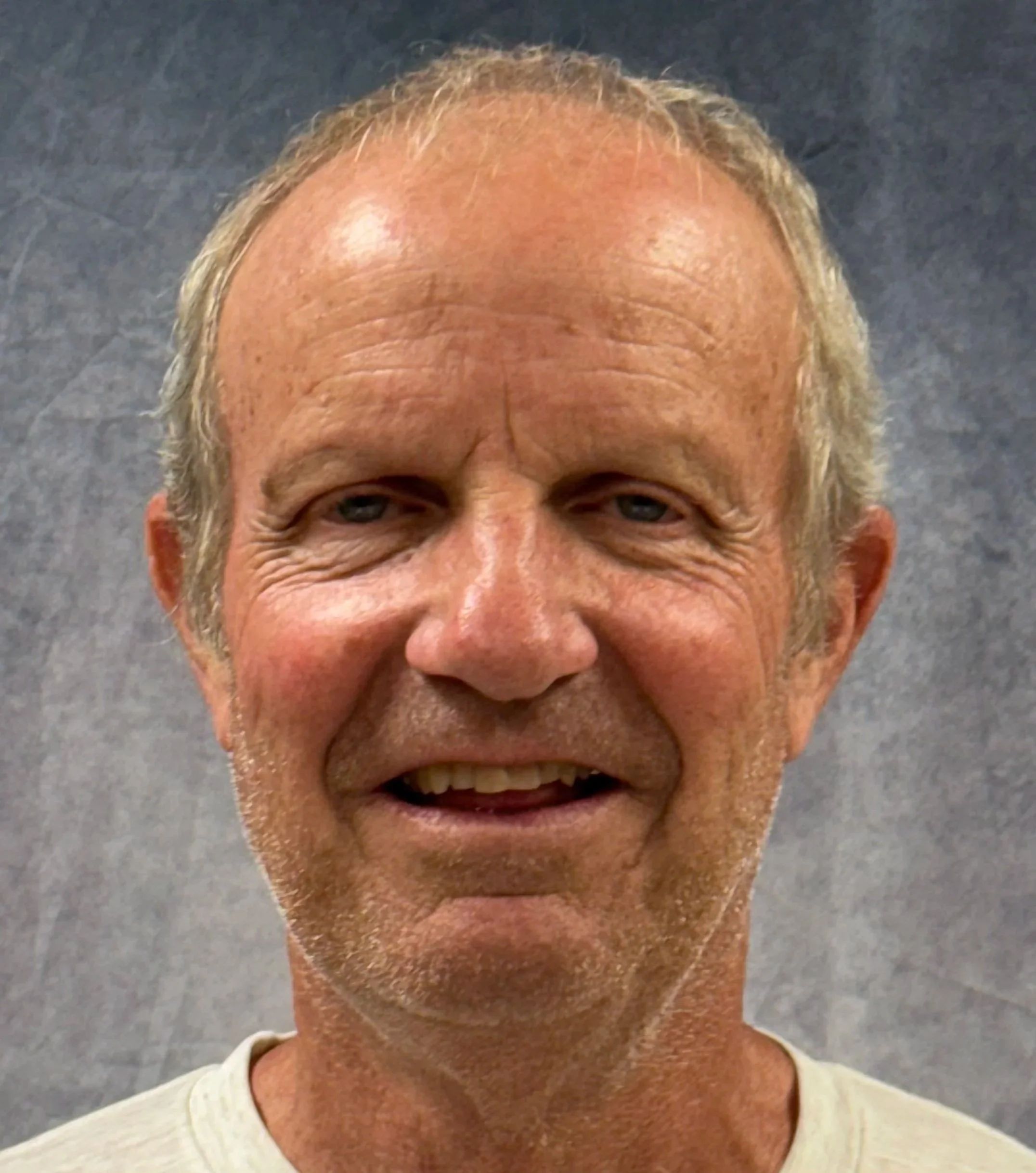 Close-up of an elderly man's face with a big smile, wearing a beige shirt and standing against a dark background.