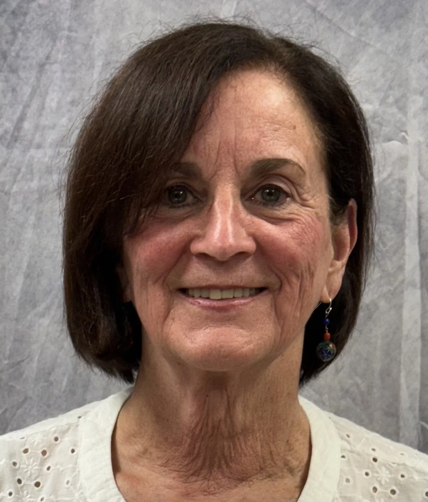 Close-up of a smiling woman with short brown hair, wearing earrings and a white eyelet blouse, in front of a gray textured background.