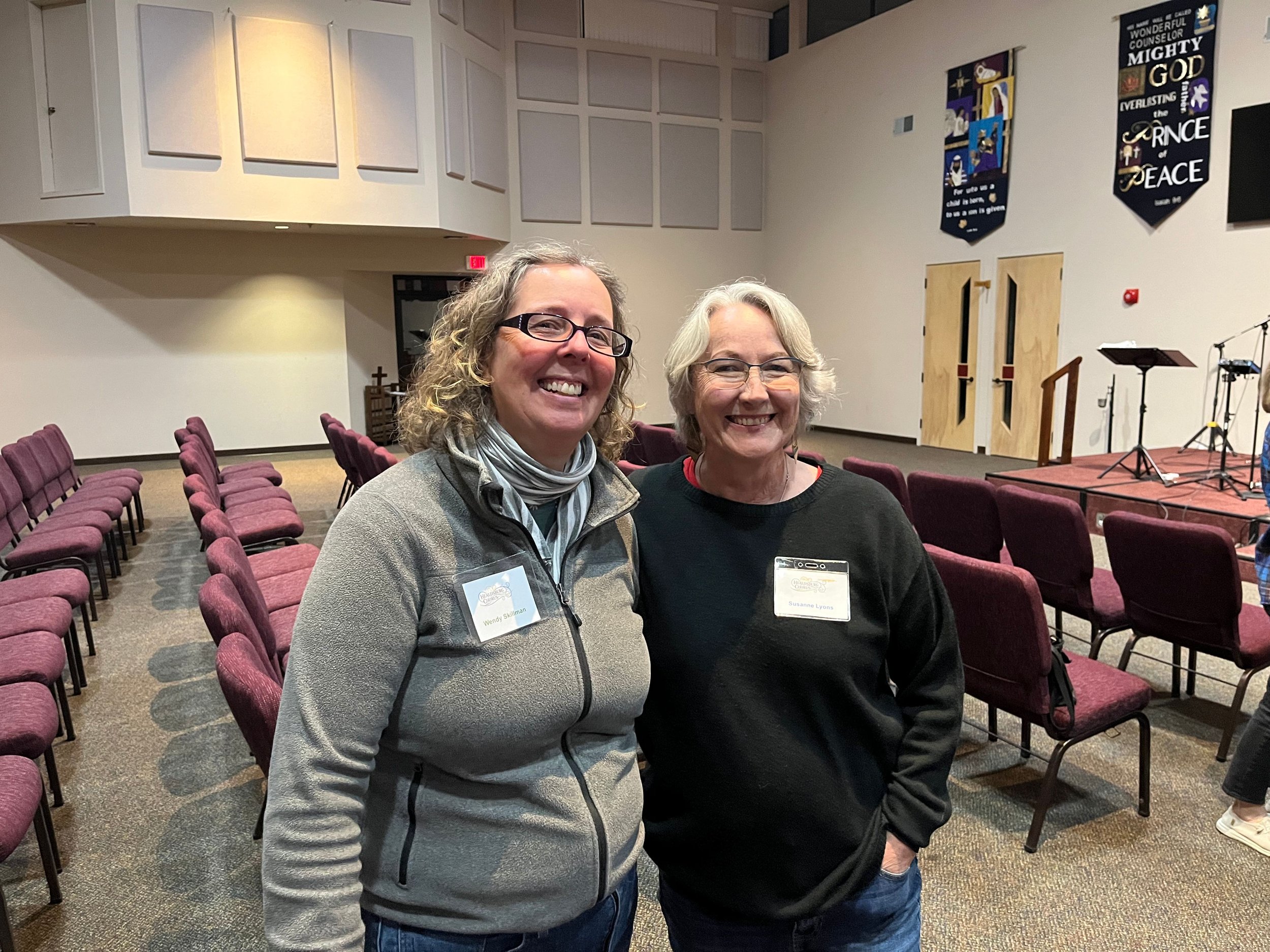 Two smiling women, Wendy Skillman and Susanne Lyons, standing inside a church or auditorium with rows of empty maroon chairs and banners on the wall, one with a cross and religious messages.