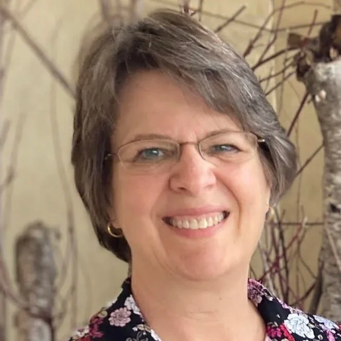 A smiling woman with short hair, glasses, and a floral blouse standing in front of a nature background.