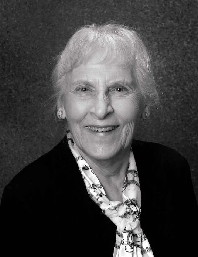 Black and white portrait of an elderly woman with short hair, smiling, wearing a dark blazer and a patterned scarf, against a dark textured background.
