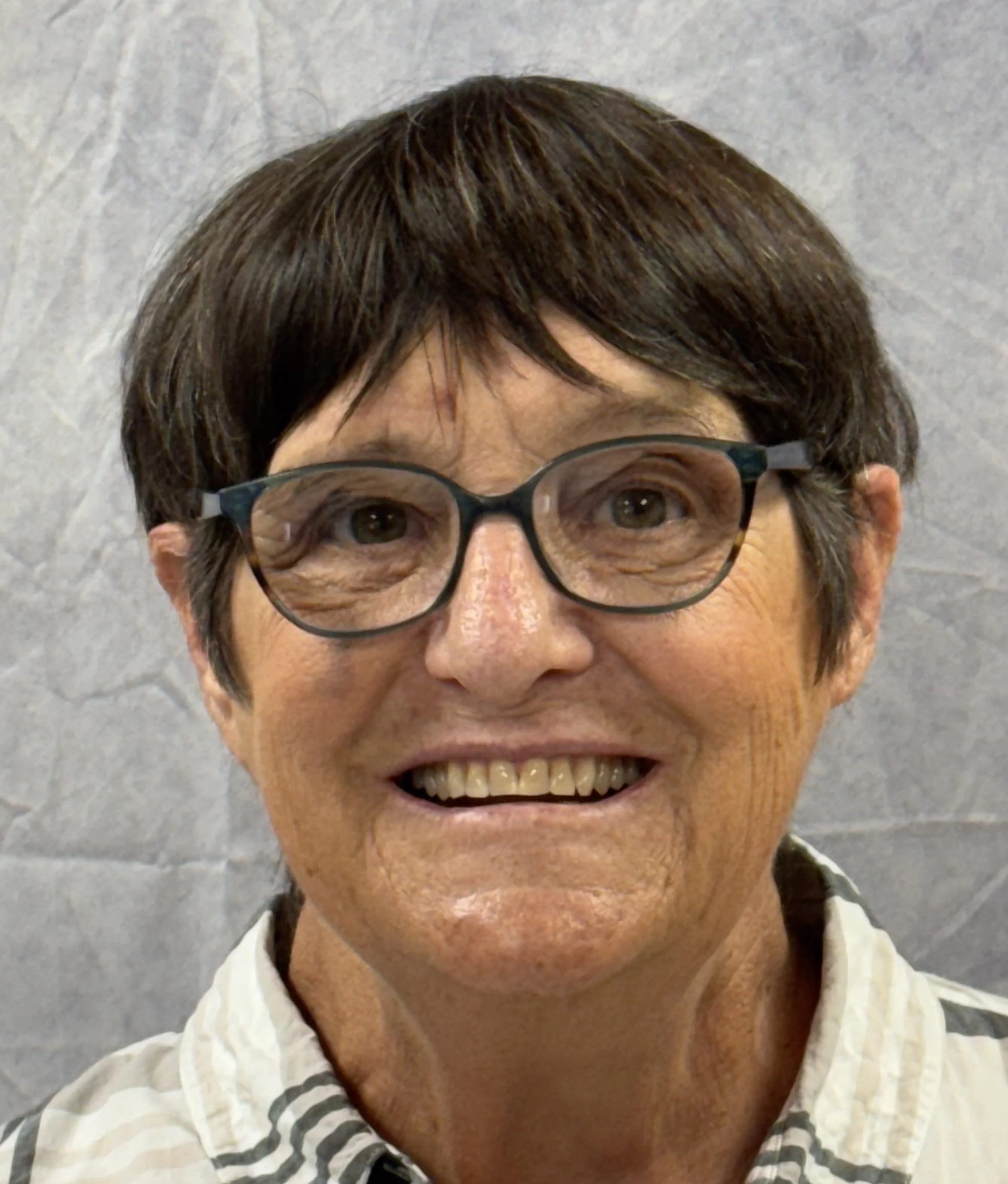 Close-up of a woman with short brown hair, glasses, and a smile, wearing a striped shirt and a white jacket, standing outdoors on a cloudy day.