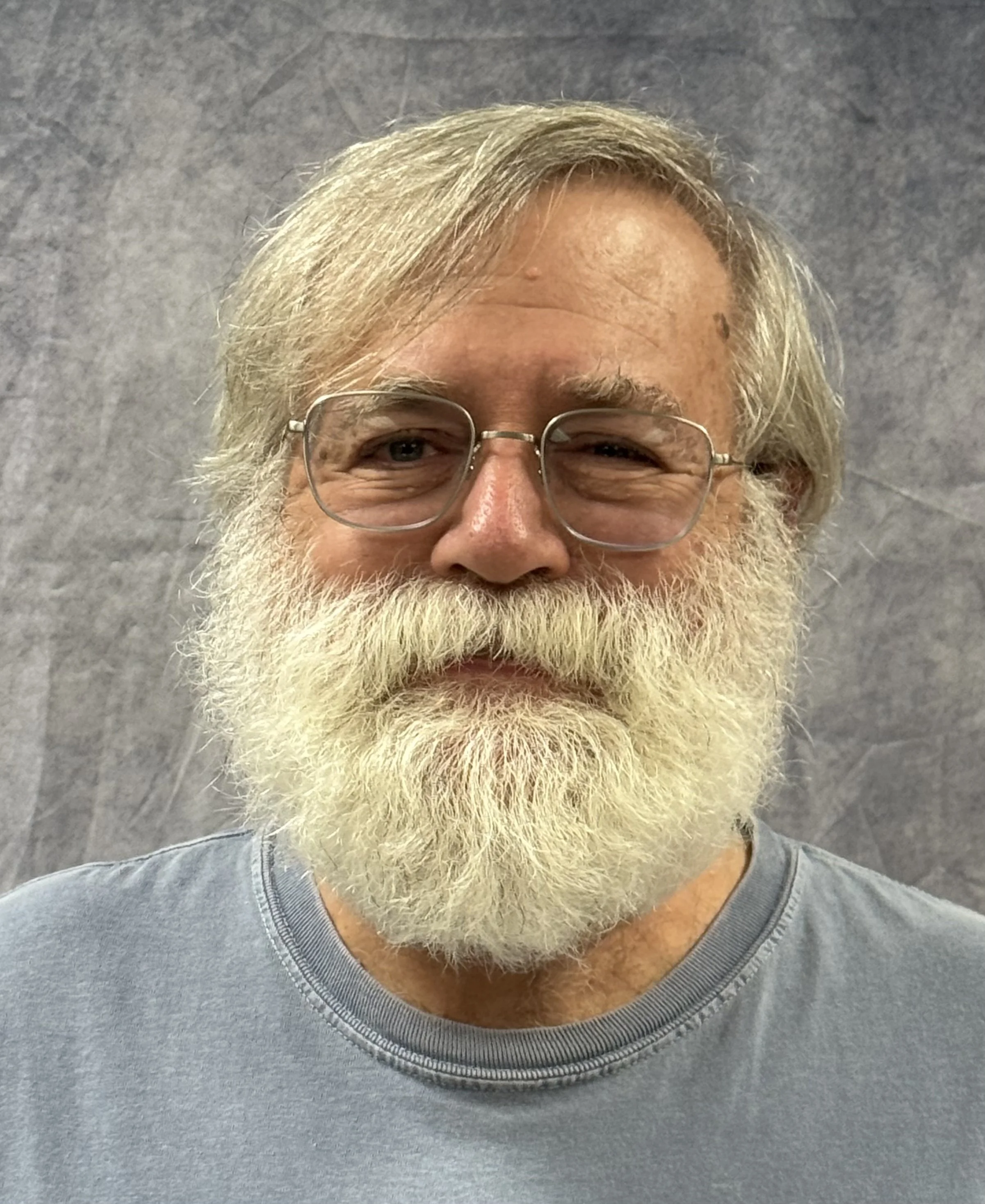 Close-up of a smiling man wearing glasses and a gray T-shirt, with gray hair and a full beard, against a gray, textured background.