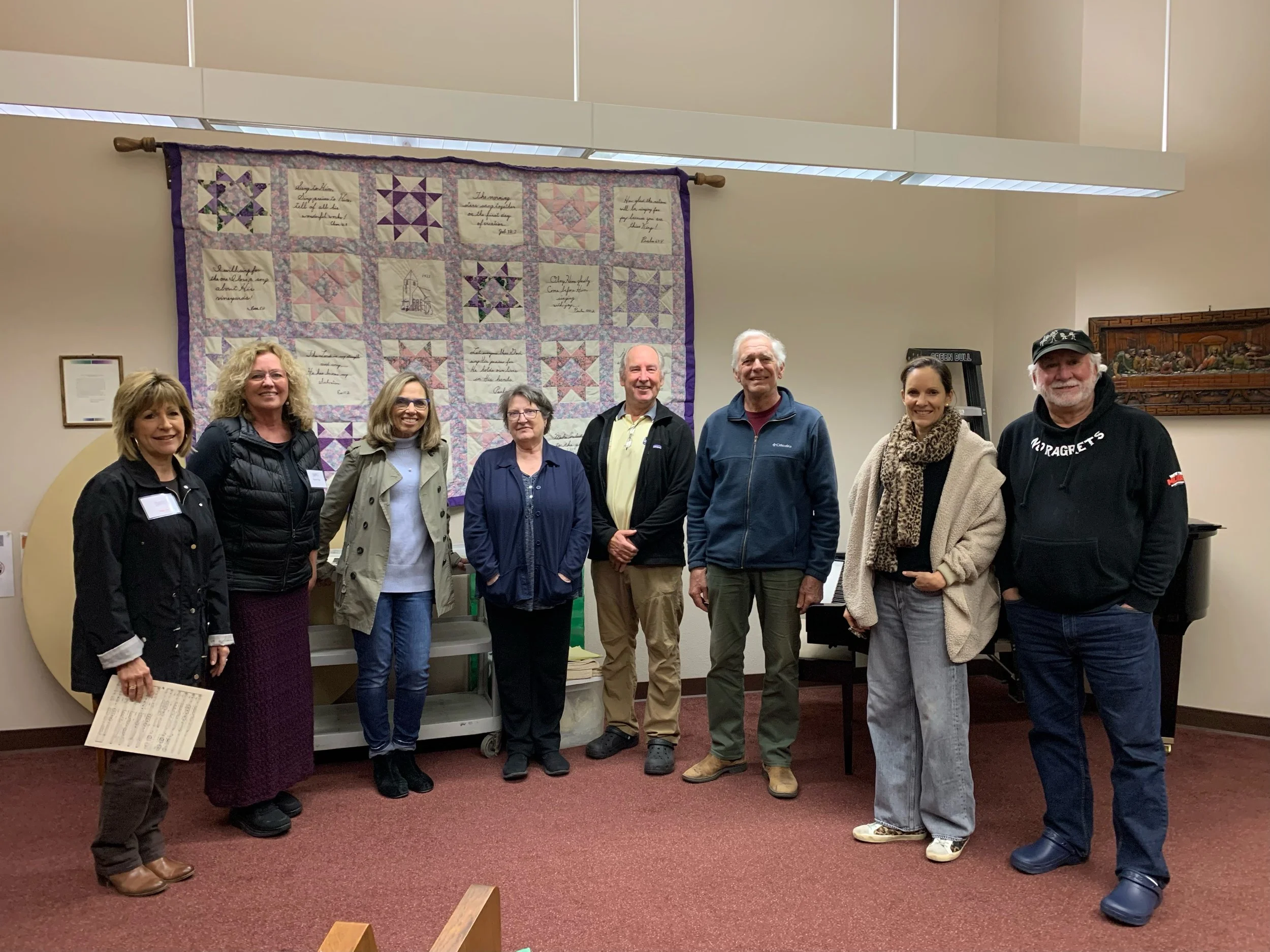 A group of eight people standing indoors in front of a wall with a quilt hanging on it. The quilt features quilt blocks and embroidered text. The individuals are smiling and dressed casually.