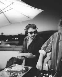 Two people at a social gathering outdoors at night, one wearing sunglasses and a jacket, the other partially visible.