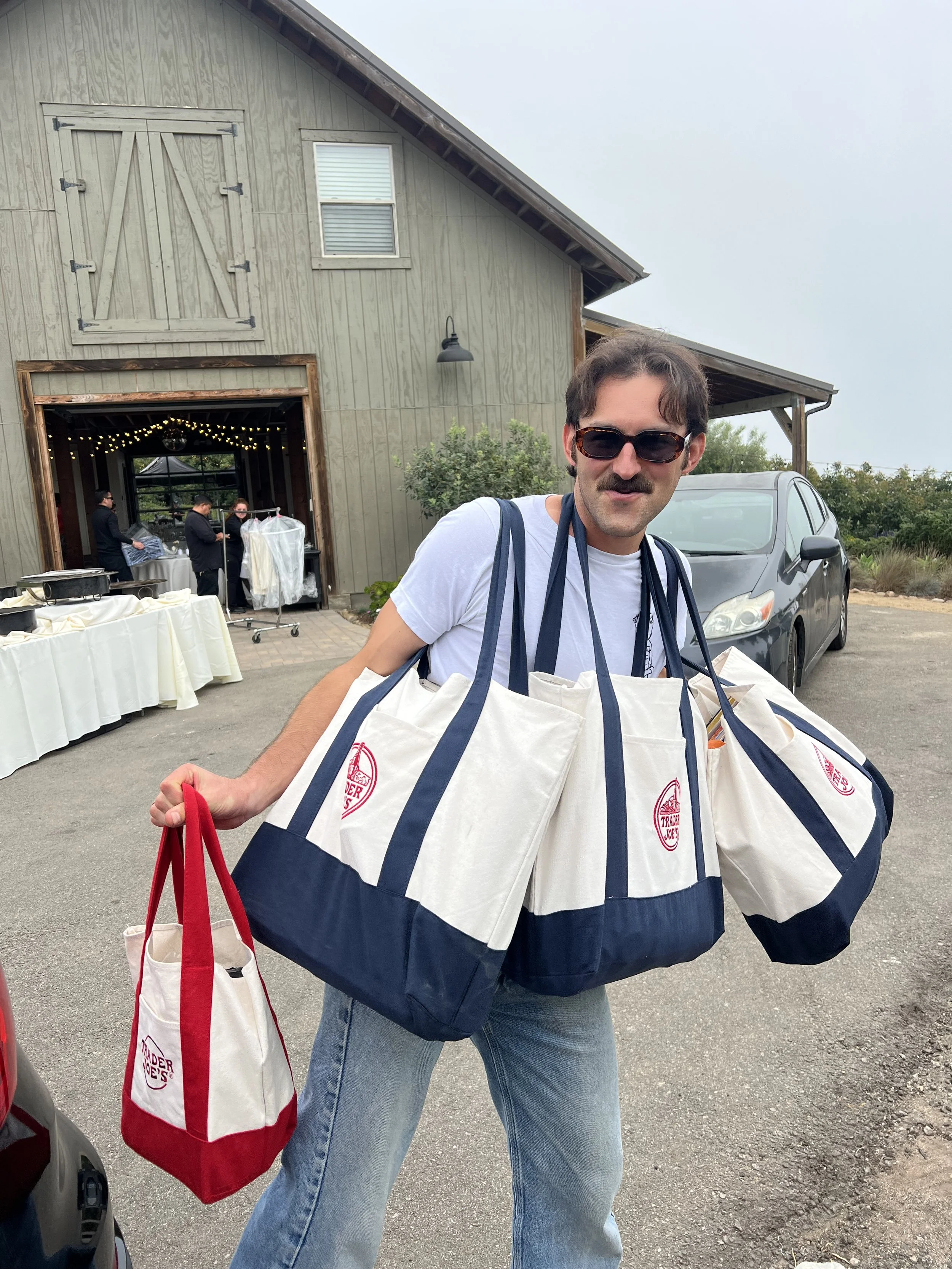 A man with sunglasses, a mustache, and dark hair holding multiple white shopping bags with red and navy blue accents outside a rustic-style building with a barn door and string lights. There are other people in the background near a table with a whit
