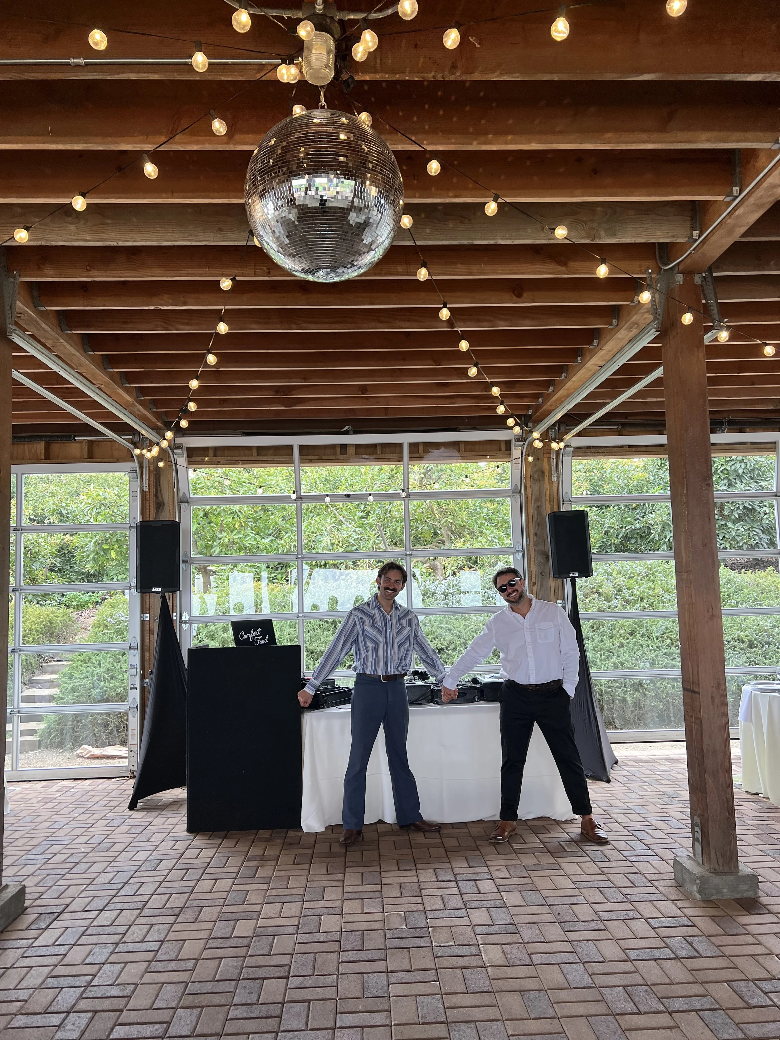 Two men are standing and smiling in front of a DJ setup inside a spacious, wooden-ceilinged venue with large windows, hanging string lights, a disco ball, and a brick floor.