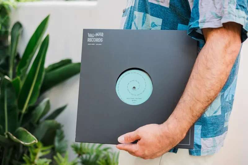 Person holding a vinyl record in a sleeve with a light blue label, standing next to green houseplants.
