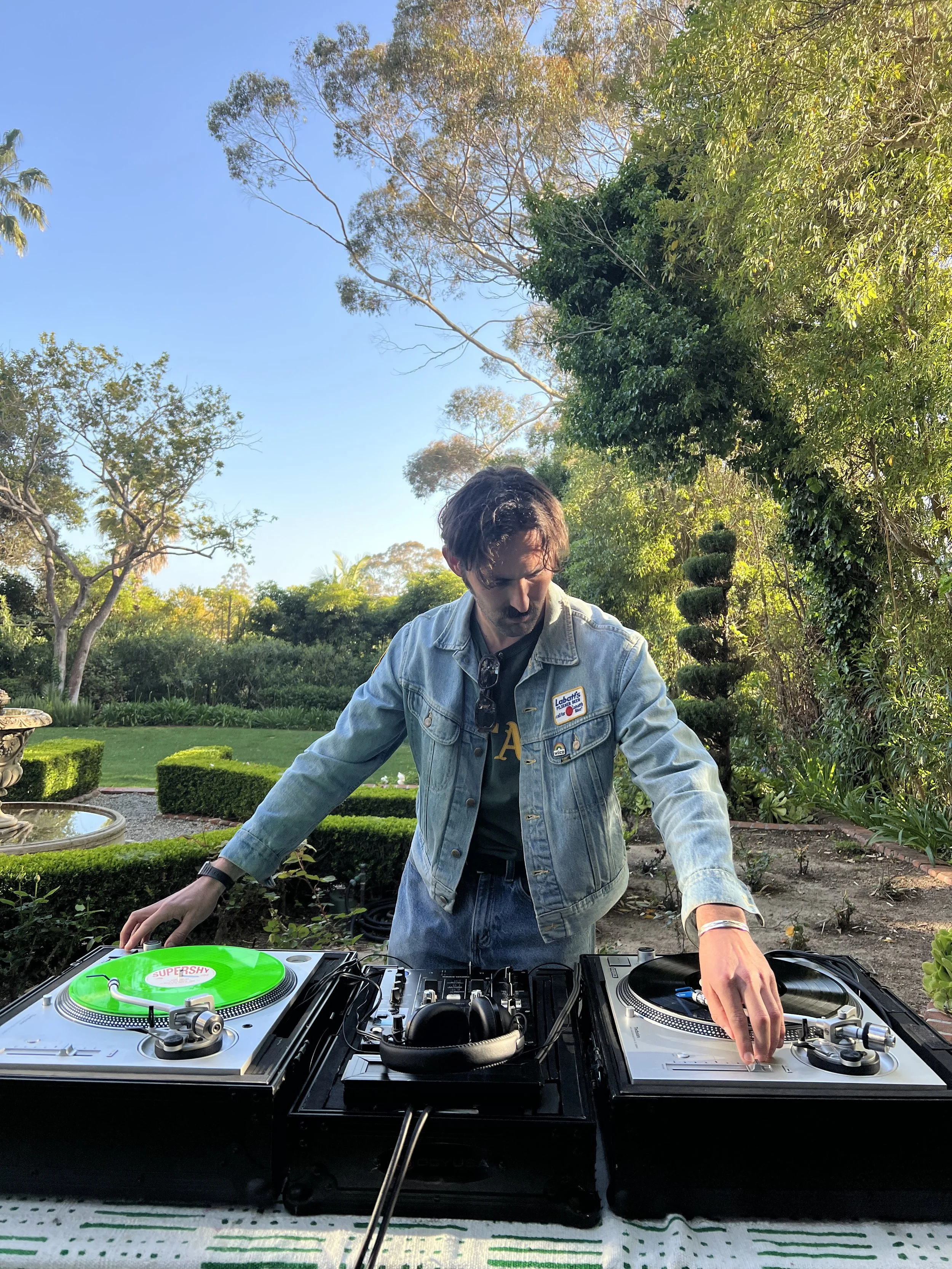 A person with brown hair wearing a denim jacket and DJ headphones is DJing outdoors surrounded by trees and greenery, with turntables and a mixer on the table.