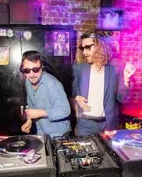 Two men DJing at a club with purple lighting, one wearing sunglasses and headphones, the other with long hair and sunglasses, in front of DJ equipment.