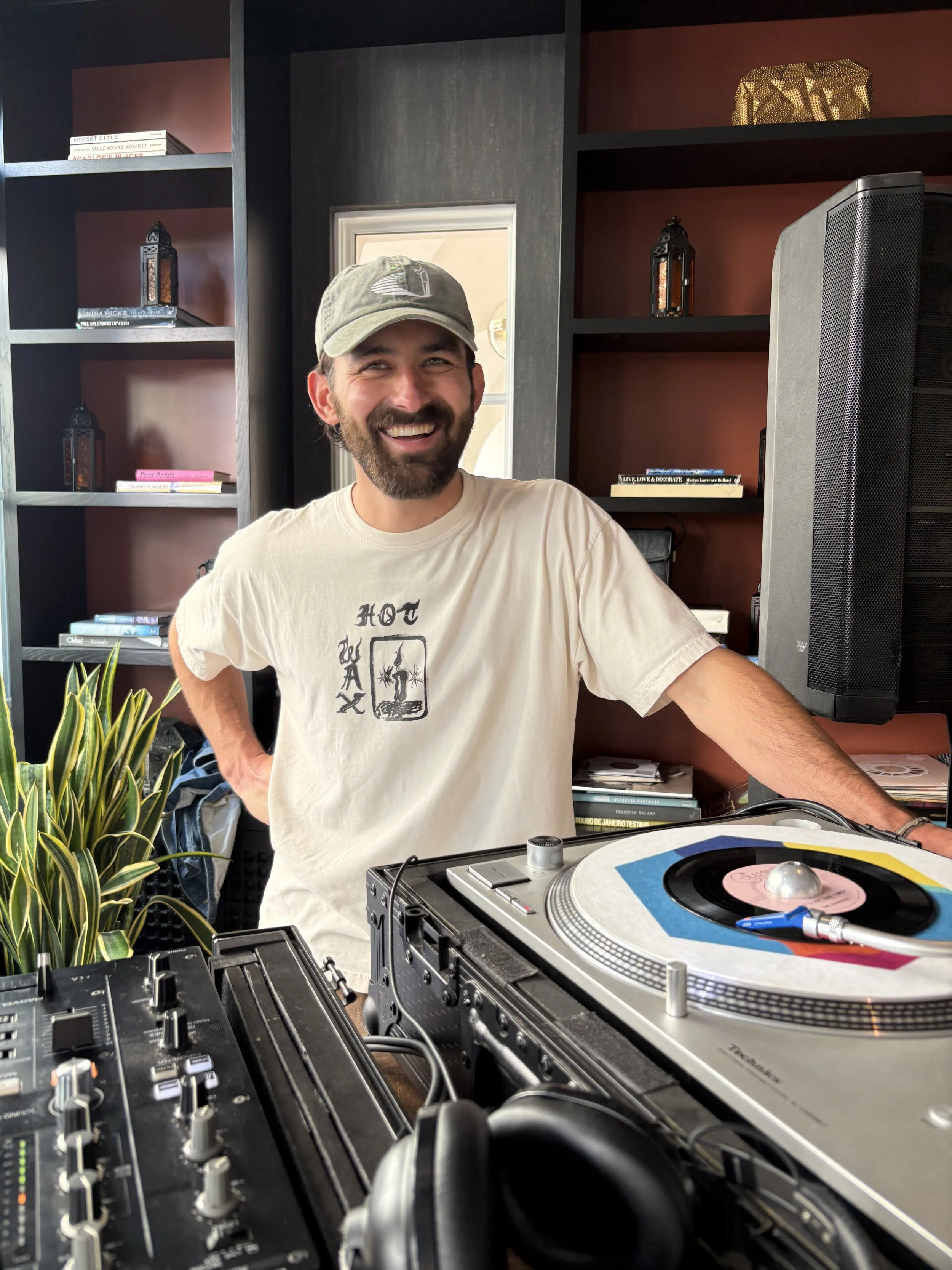 Smiling man with a beard wearing a beige t-shirt and cap, standing behind DJ equipment with turntables and a mixer, in a room with shelves holding books, lanterns, and a plant.