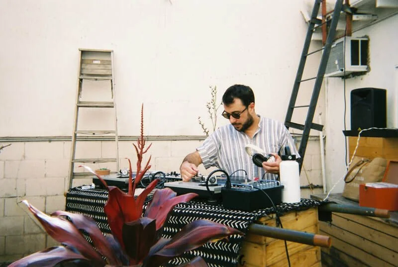 A man with sunglasses wearing a striped shirt, working with DJ equipment outdoors.