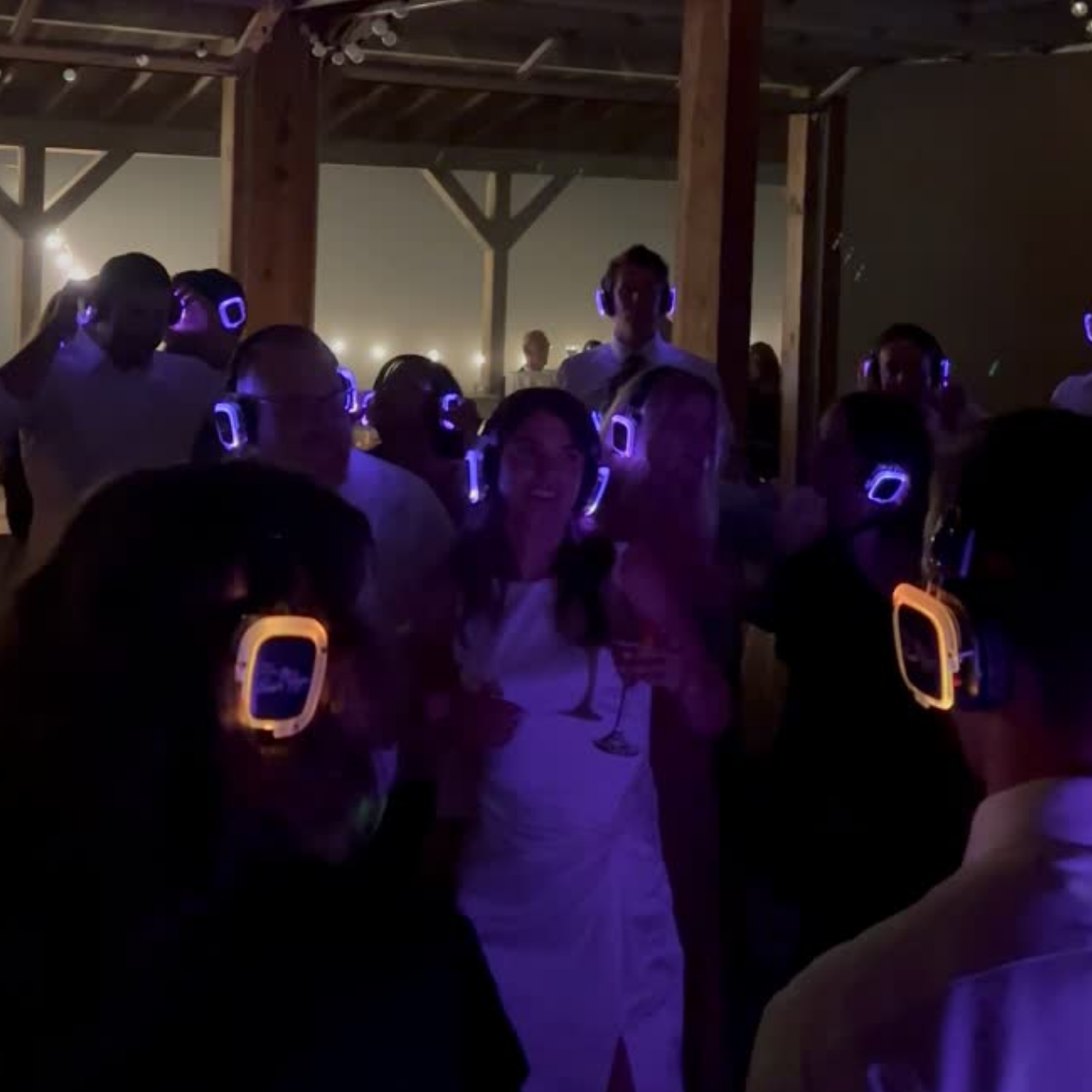 Group of people wearing glowing blue LED headphones at an indoor event, with wooden beams and minimal lighting in the background.