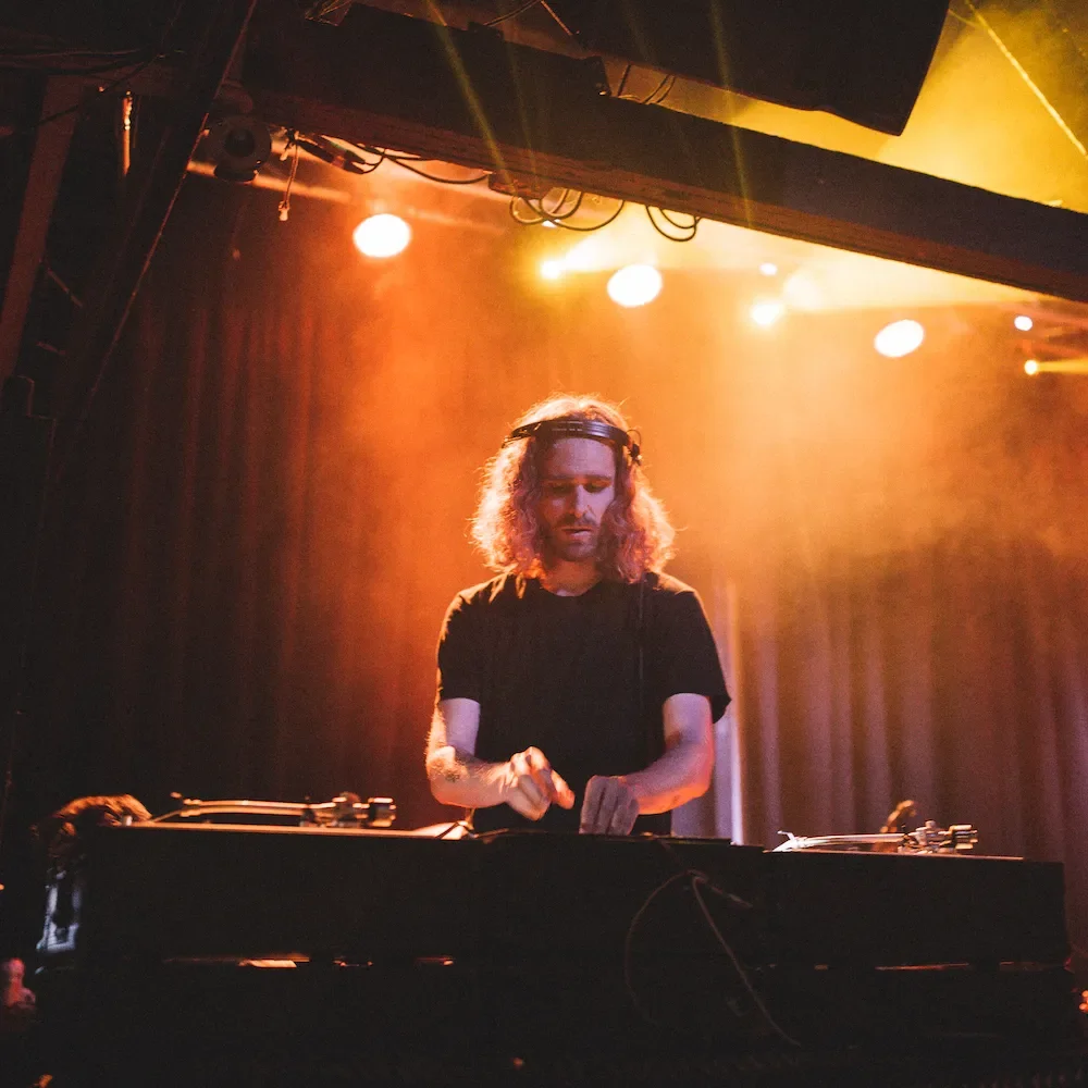 A DJ with long curly hair wearing headphones, working behind a turntable in a dimly lit nightclub or concert venue with orange and yellow lighting.